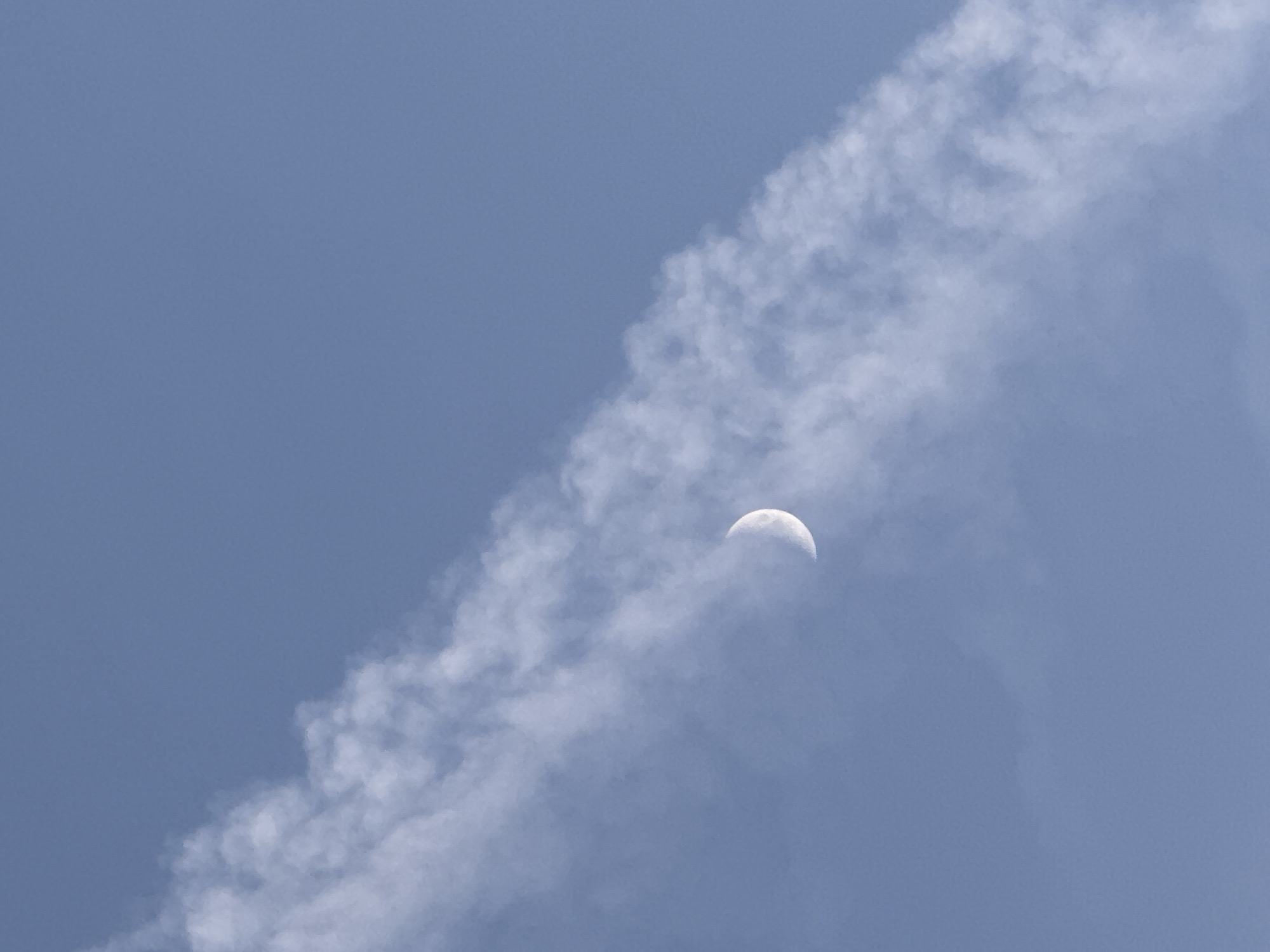 The image shows a half-moon in a light blue sky, partially obscured by a wispy, diagonal band of clouds. There are no people or identifiable objects beyond the moon and clouds. The scene is serene and focused on the celestial elements. No information in the image confirms that the image was taken in Chaikovsky, Russia.
