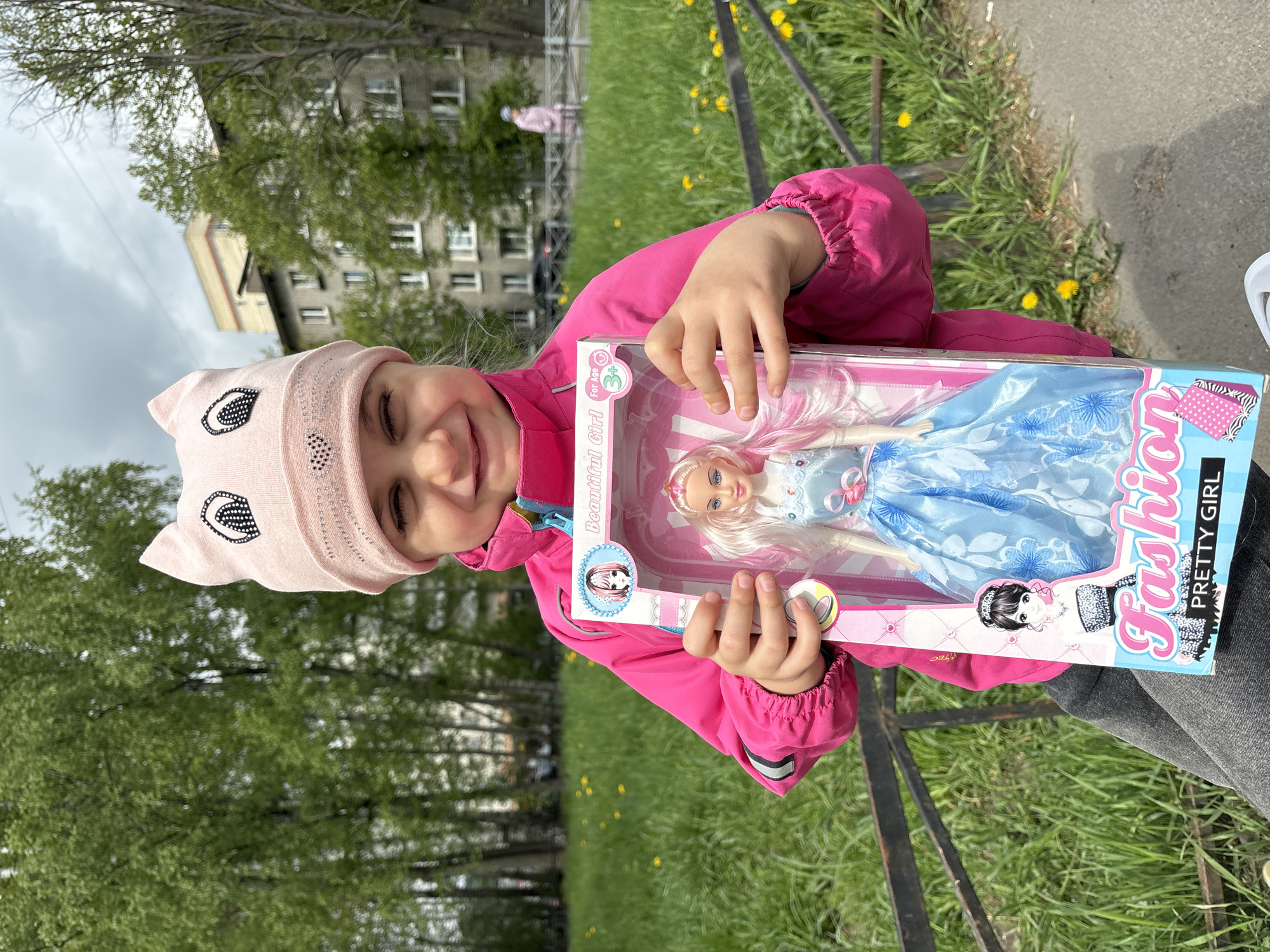 A young girl in Saint Petersburg, Russia, is holding a boxed fashion doll. She is wearing a pink jacket and hat, and she smiles at the camera. The doll is dressed in a blue gown. The scene appears to be outside, near a grassy area with dandelions. In the background is a residential building, indicating an urban setting.