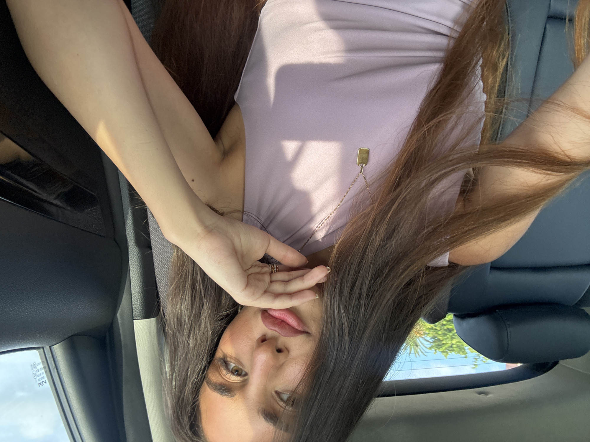 A woman with long brown hair is in the driver's seat of a car. She is wearing a light purple top and a gold necklace. Her right hand is near her face. The photo appears to be taken from inside the vehicle. The exterior environment is visible through the car window. The city of Boca Raton, United States of America, cannot be verified from the image.