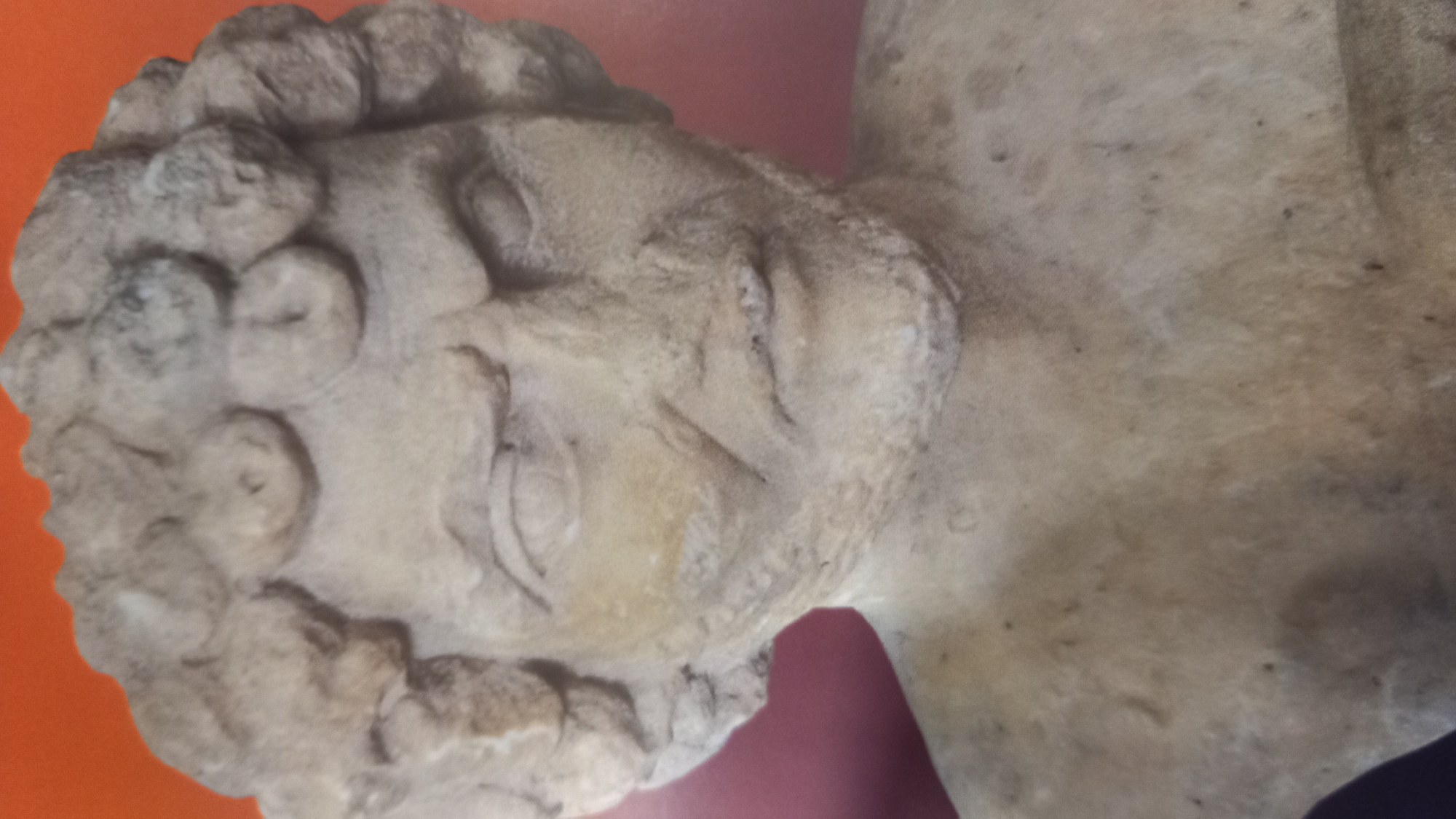 The image is a close-up, head-and-shoulders shot of a weathered, beige stone bust of a male figure. The bust is positioned against a vibrant orange background. The figure has a stern expression, with furrowed brows and intense, slightly downward-cast eyes. His hair is depicted as a series of thick, curly locks arranged around his head. He has a prominent nose, a small, pursed mouth, and a rough, textured beard. The stone shows signs of age and erosion, with visible pitting and some discoloration. The overall impression is of an ancient artifact, likely a statue or relief. This bust is located in the Archaeological Museum of Kos, in the city of Kos, Greece.