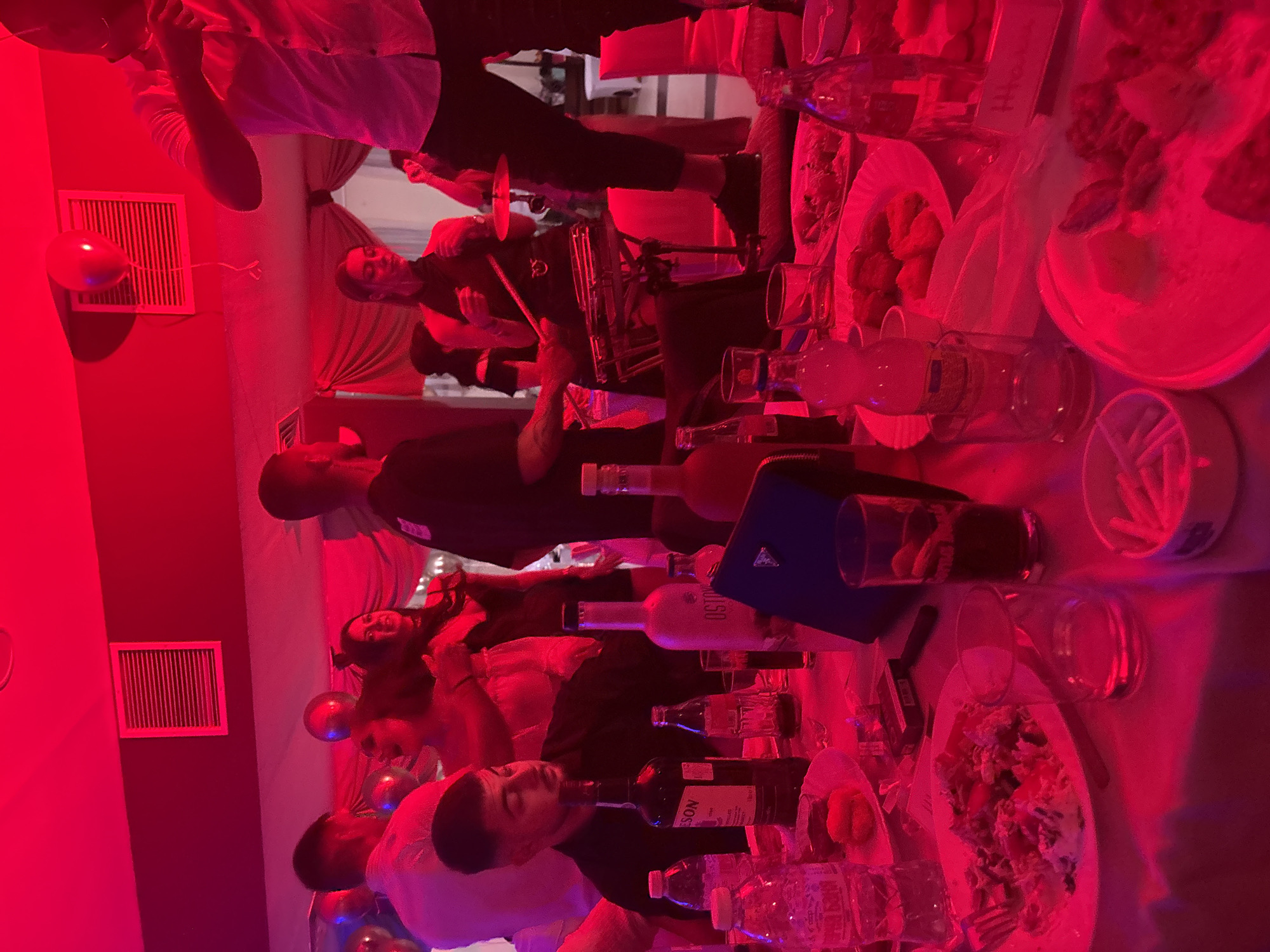 The image depicts an event or gathering in what appears to be a restaurant setting, potentially in Kostinbrod, Bulgaria. A drummer is playing an instrument, seemingly providing entertainment for the occasion. Several people are present, some standing and some seated around tables filled with food, drinks, and bottles. The overall environment seems lively and social. The dominant red lighting makes it difficult to discern specific details about the individuals or the environment.