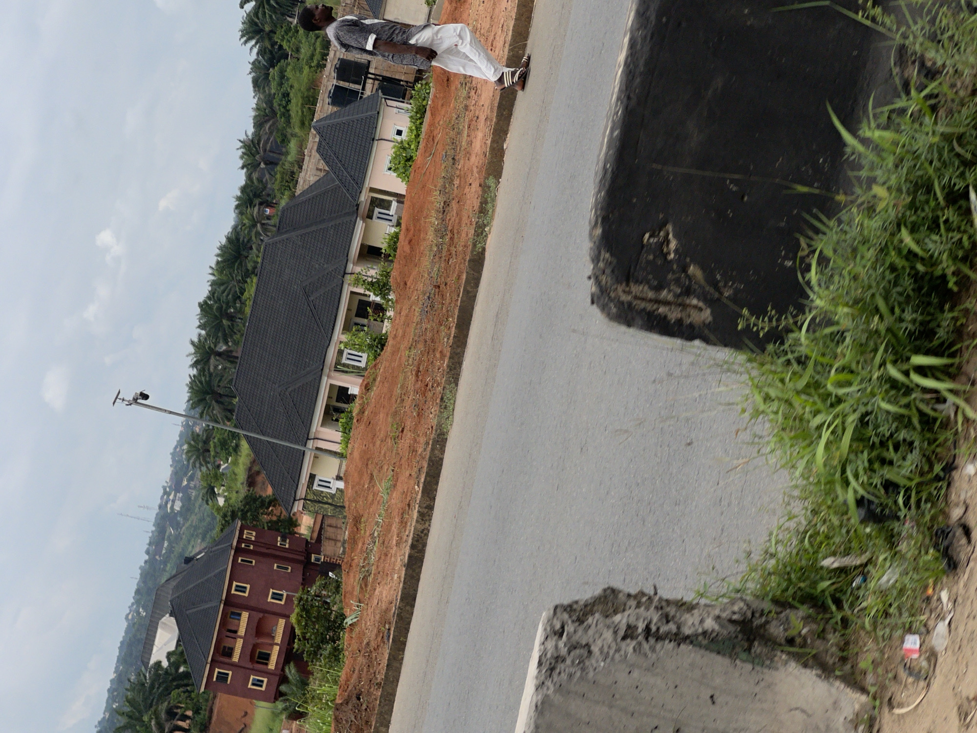 An overcast, medium-shot, eye-level, outdoor photograph captures a man walking on a paved road. The man, dressed in a grey and white patterned shirt and white pants, walks away from the camera towards the right side of the frame. He is wearing sandals and has dark skin and hair. In the background, a cluster of modern, multi-story buildings with dark roofs are visible, some appearing to be under construction. Palm trees and other vegetation dot the landscape, which includes rolling hills. A security camera is mounted on a tall pole near the buildings. The foreground is dominated by a gray concrete barrier on the left and a rough, dark barrier on the right, both partially obscuring the road. Lush green grass grows along the side of the road and around the concrete barriers, with some scattered debris visible. The scene is located in Obosi, Nigeria.