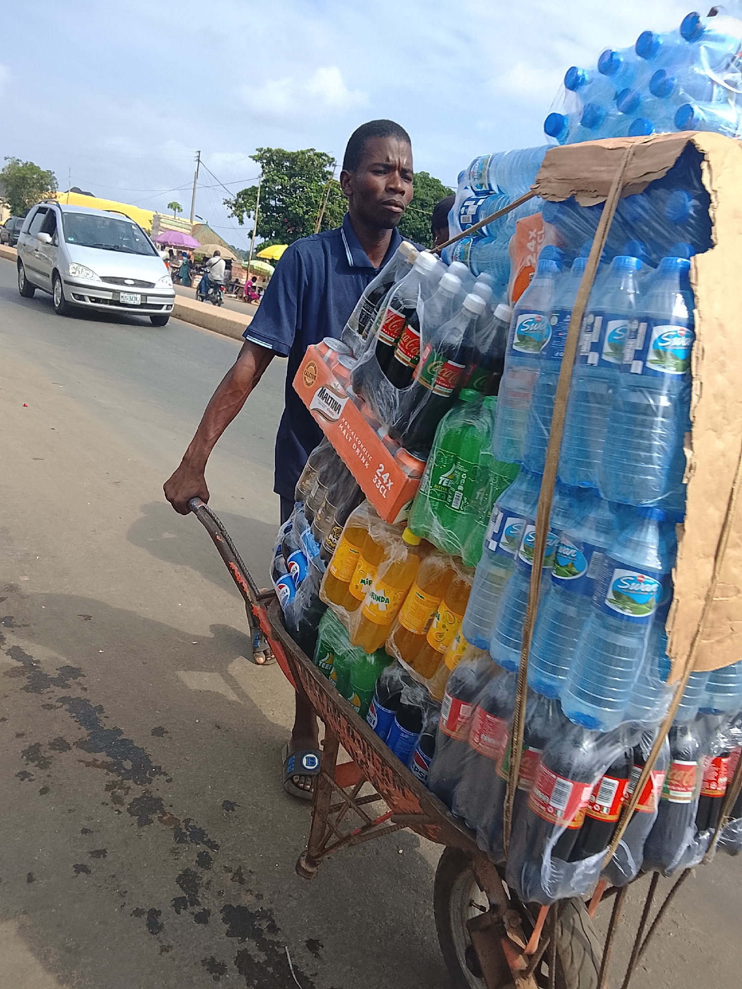In Akwanga, Nigeria, a man pushes a wheelbarrow laden with bottled drinks. The wheelbarrow, made of red metal with a single wheel, is overloaded with various beverages. Dominating the load are stacks of blue-bottled water, some with green labels. Interspersed among the water bottles are cases of brown sodas, some with red Coca-Cola branding and others with blue Pepsi branding. There are also yellow-bottled drinks, possibly orange juice or soda, and green-bottled beverages. A brown cardboard box, possibly for packaging, is propped up against the bottles. The man, dressed in a navy blue collared shirt, wears sandals and appears to be walking on an asphalt road. In the background, a silver SUV is visible, along with other vehicles and pedestrians on a street lined with trees and buildings. The sky is overcast with scattered clouds.