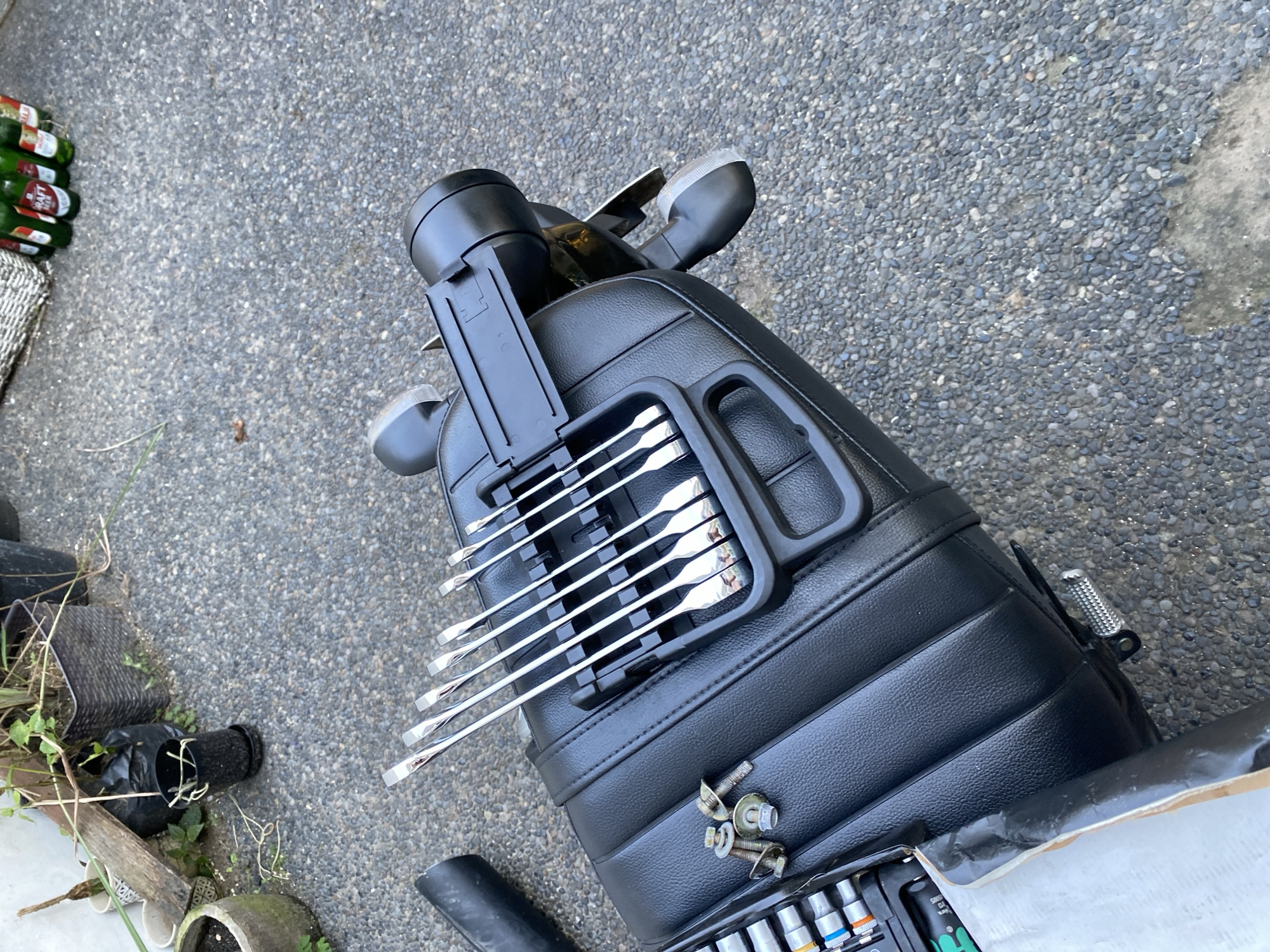 A motorcycle seat with tools and hardware rests on a gravelly surface in Kuta Selatan, Indonesia. A set of flathead screwdrivers is neatly organized in a black plastic holder secured to the seat. To the left of the screwdrivers, a toolbox containing various sockets and a few loose screws and bolts is partially visible. A black pipe segment is positioned to the left of the seat. In the background, potted plants and plastic bags suggest an outdoor setting. Several green beer bottles are scattered on the ground in the upper right corner. The lighting indicates it is daytime.