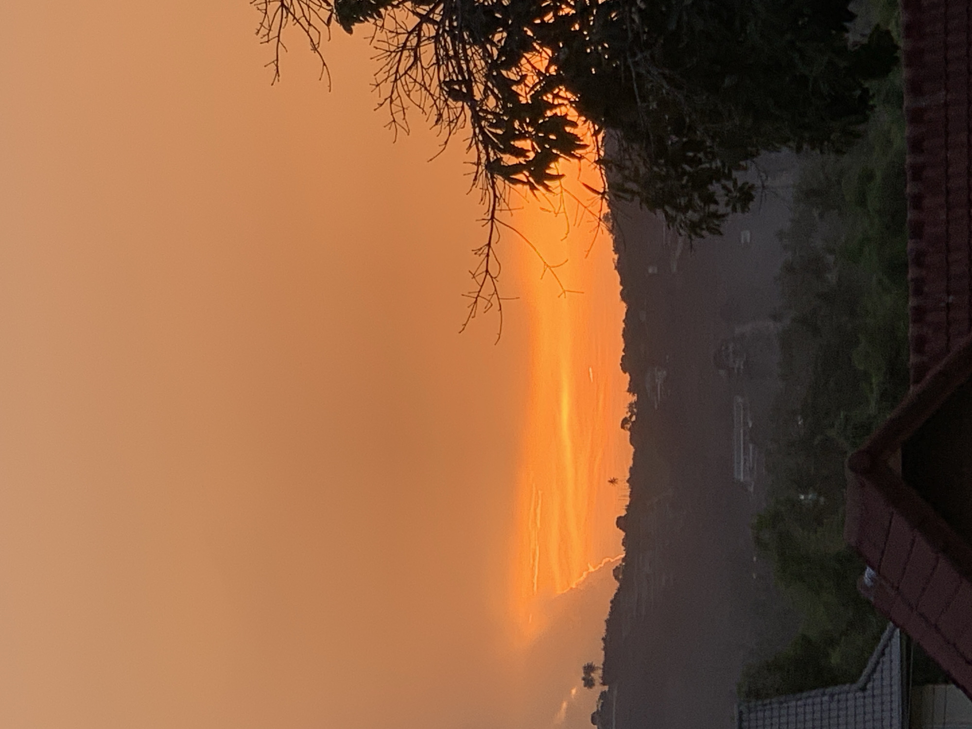 The image displays a stunning sunset or sunrise over a natural landscape, viewed from an elevated vantage point. The sky is the dominant feature, painted with vibrant hues of orange and golden yellow, brightest along the horizon and fading into a softer, dusky orange towards the top left. Streaks of light and cloud formations are visible within the fiery portion of the sky.
A dark, silhouetted line of hills or mountains stretches across the middle of the frame, delineating the horizon against the colorful sky. In the immediate foreground, the intricate silhouettes of tree branches, some bare and some adorned with leaves, frame the upper left and right portions of the image. On the far right, part of a building with a reddish-brown tiled roof is visible, also in silhouette, suggesting a structure near the viewpoint.
The scene evokes a sense of tranquility and natural beauty, characteristic of dusk or dawn. There are no visible people or discernible activities taking place beyond the natural progression of day into night or vice versa. No text is visible in the image. The location context for this picturesque view is บ้านบางรักษ์, Thailand.