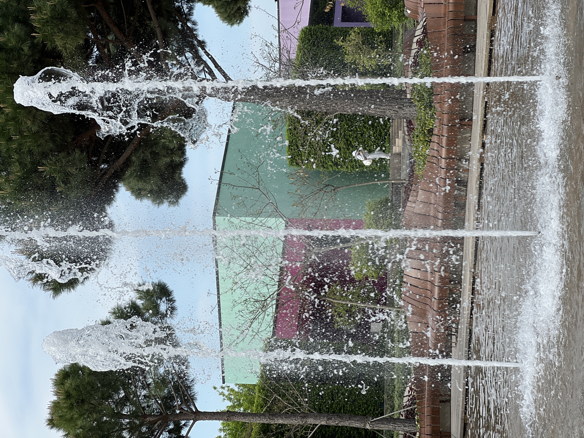 The image shows a fountain with multiple streams of water shooting upwards. Behind the fountain, trees and a building with a light green and maroon facade are visible. The location is Krasnodar, Russia. A figure in white is walking on a walkway or steps, partially obscured by greenery.