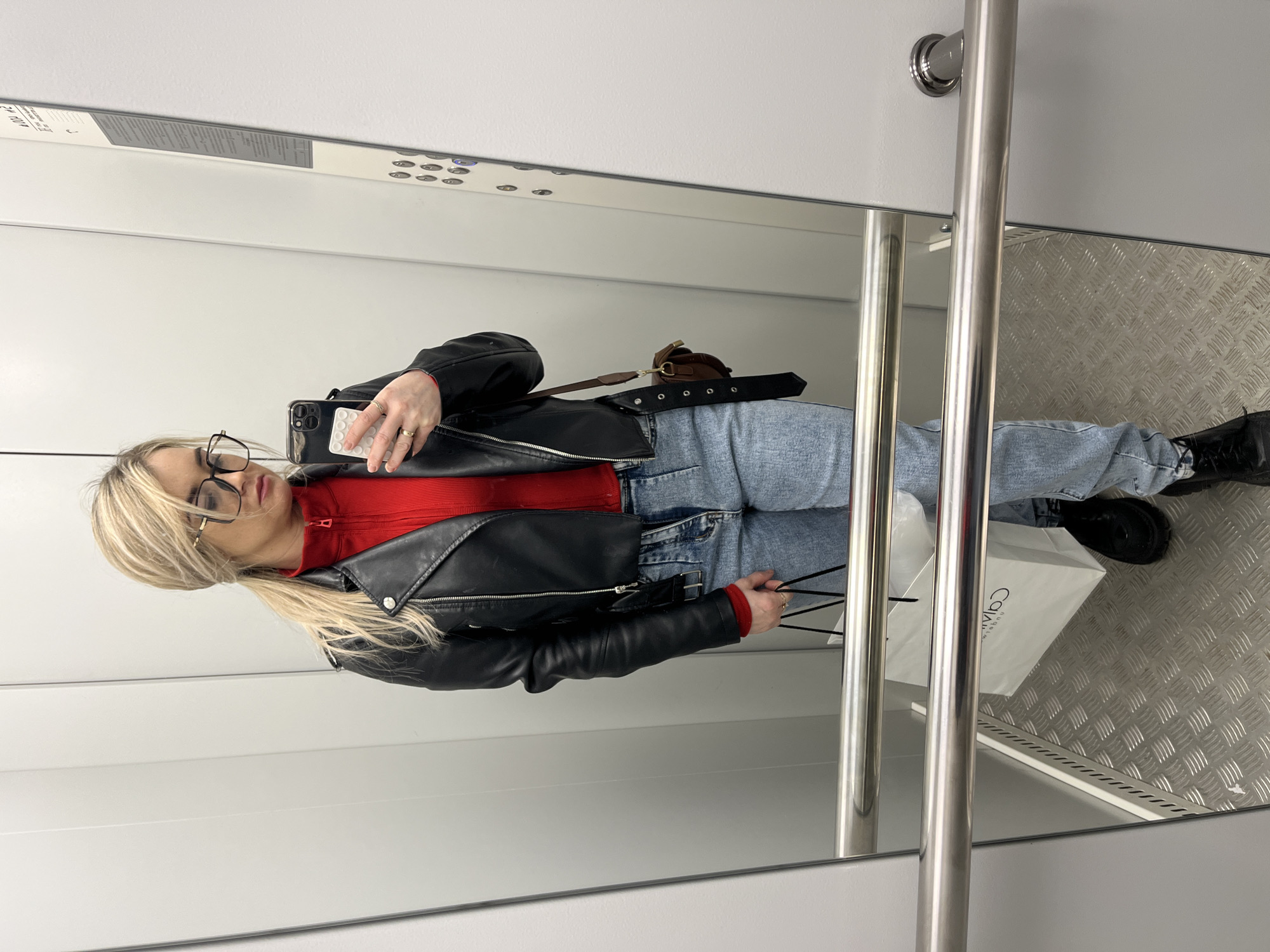 The image depicts a woman standing in an elevator. She is wearing a red top, light blue jeans, and a black leather jacket. She has blonde hair pulled back in a ponytail and is wearing glasses. She is holding a phone in one hand and a white shopping bag, presumably from Calvin Klein, in the other. A brown belt is visible, placed on top of the jeans. The elevator has stainless steel handrails and a patterned metal floor. The walls are light-colored, possibly white or light grey, with a panel of elevator buttons visible in the background. The location is likely Chaikovsky, Russia, as requested. There is no discernible action or interaction occurring beyond the woman standing for the photo.