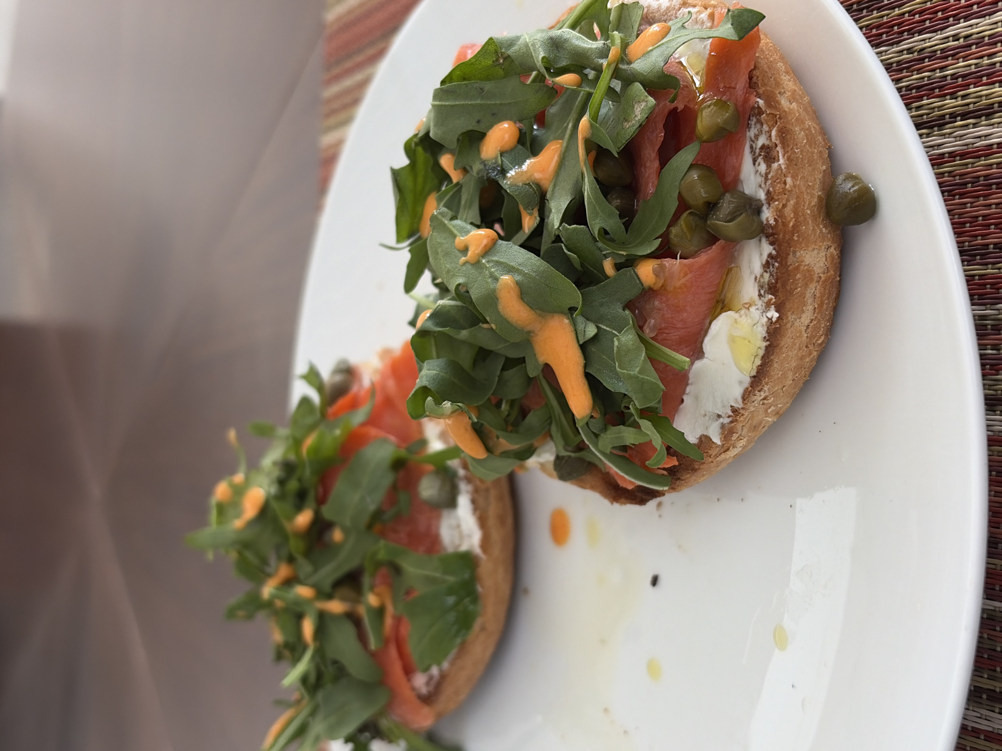 The image shows two open-faced sandwiches arranged on a white plate. Each sandwich consists of a bun, a creamy white spread, sliced salmon, green leaves (likely arugula), capers, and a drizzled orange sauce. The plate is placed on a textured surface resembling a woven placemat. The background is blurred, but a neutral-toned wall is visible. The scene is lit with natural light. There is no indication of the city of San Bernardino Tlaxcalancingo, Mexico.