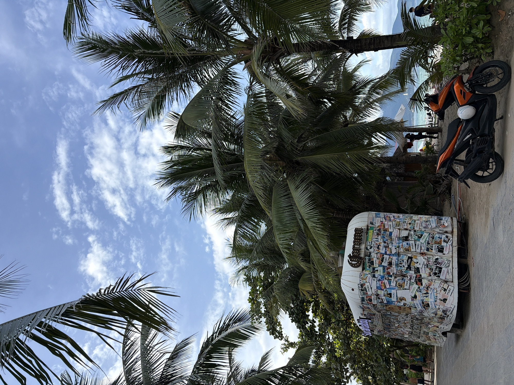 The image depicts a beachside scene in Đà Nẵng, Vietnam, featuring lush palm trees framing a bright blue sky with scattered white clouds. In the foreground, an orange scooter is parked on a sandy path, a white helmet resting on its seat. Behind the scooter, a white food truck is covered in numerous colorful stickers and signage, with a prominent "Corona" logo at the top. Several people, casually dressed, are visible walking along the path and in the distance, near the beach. Further back, the ocean meets a distant coastline with mountains under a hazy sky. The overall atmosphere is that of a tropical vacation spot.
