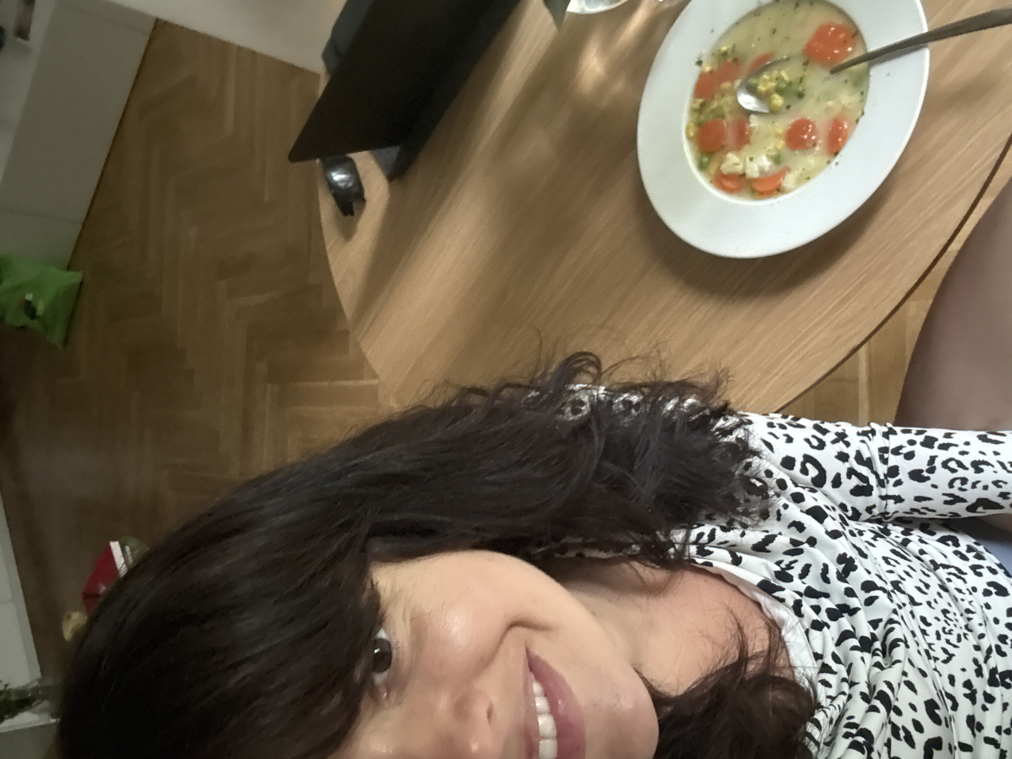 A woman with dark hair and wearing a white shirt with black leopard print is seated at a light brown, round table. A white plate of soup, containing orange slices and a spoon, rests on the table. The table is in an interior space with a herringbone wood floor pattern. A black object, possibly a TV stand or speaker, is also present on the table. The location is not possible to determine within Berlin, Germany.