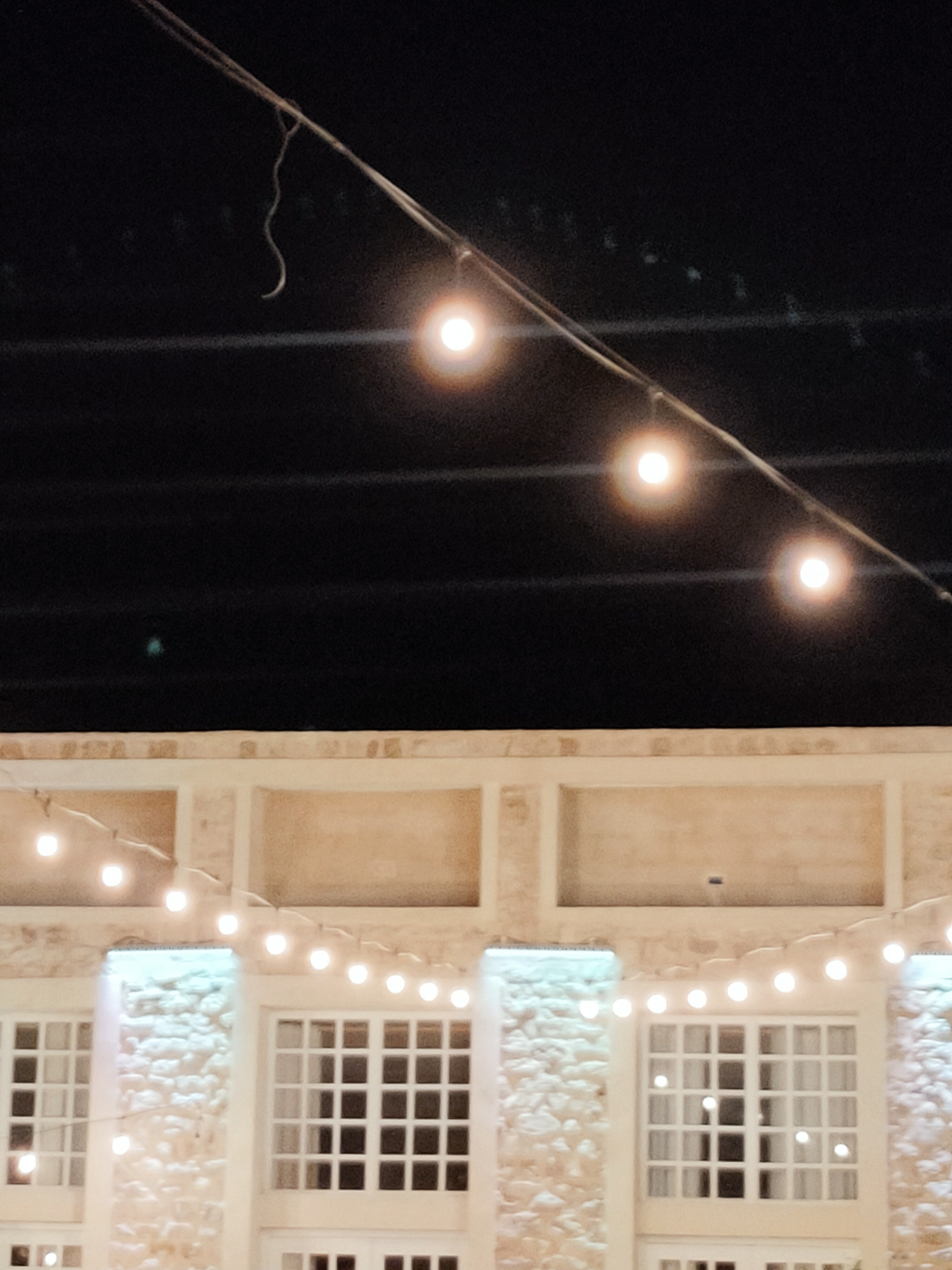 The image depicts an outdoor scene at night, possibly in Cebalat, Tunisia, featuring a building facade illuminated by string lights. The upper portion of the image shows a dark sky with a wire strung diagonally across it, supporting several glowing bulbs. A thin, curved, white object, resembling a detached vine or string, hangs from the wire near the top left. The lower part of the image reveals the facade of a building constructed with stone and plaster elements. A line of string lights runs horizontally across the facade, casting a warm glow. Below the string lights, several large windows with white frames and grid-like panes are visible. The windows are dark, suggesting they are either closed or the interior is unlit. The stone and plaster details of the building suggest a rustic or traditional architectural style. The overall impression is one of an evening setting, likely for an outdoor gathering or event, with decorative lighting enhancing the building's appearance. There are no people or animals visible in the image.