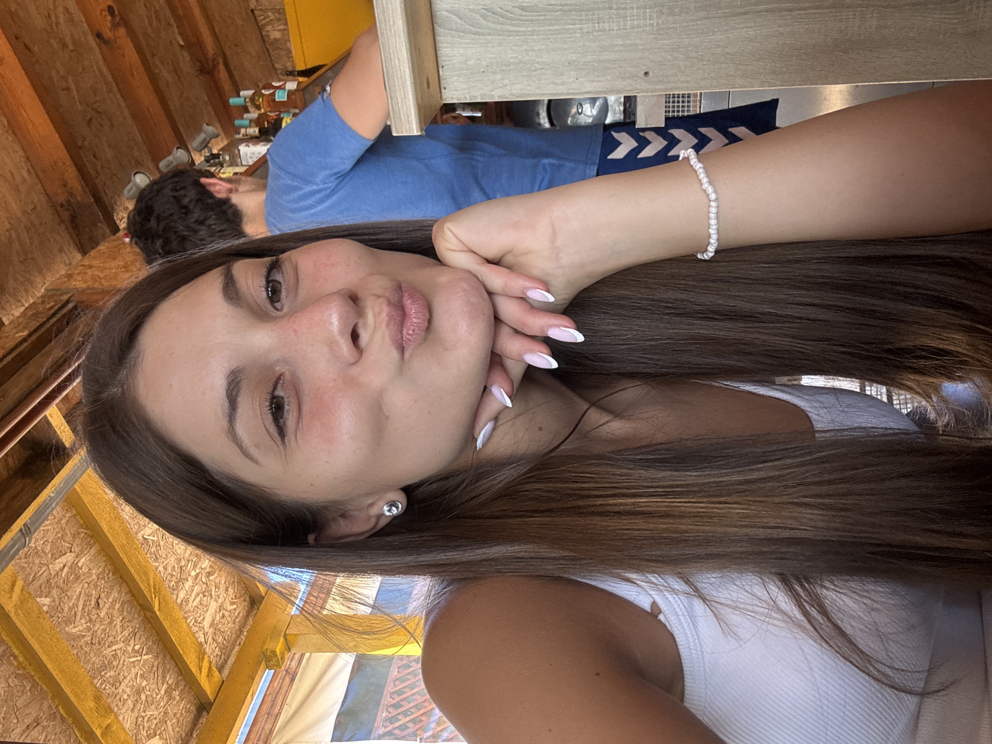 A young woman with long brown hair and a white tank top puckers her lips for a selfie. She is leaning her chin on her hand, which is adorned with a pearl bracelet and has white-tipped fingernails. A man in a blue t-shirt stands behind her in the background, near shelves stocked with bottles. The scene appears to be an outdoor bar or cafe with a wooden roof and a counter. The lighting suggests it is daytime. The woman's skin has a slight tan and a scattering of freckles. The location is the bar at "The Old Fisherman's House" restaurant in Sozopol, Bulgaria.
