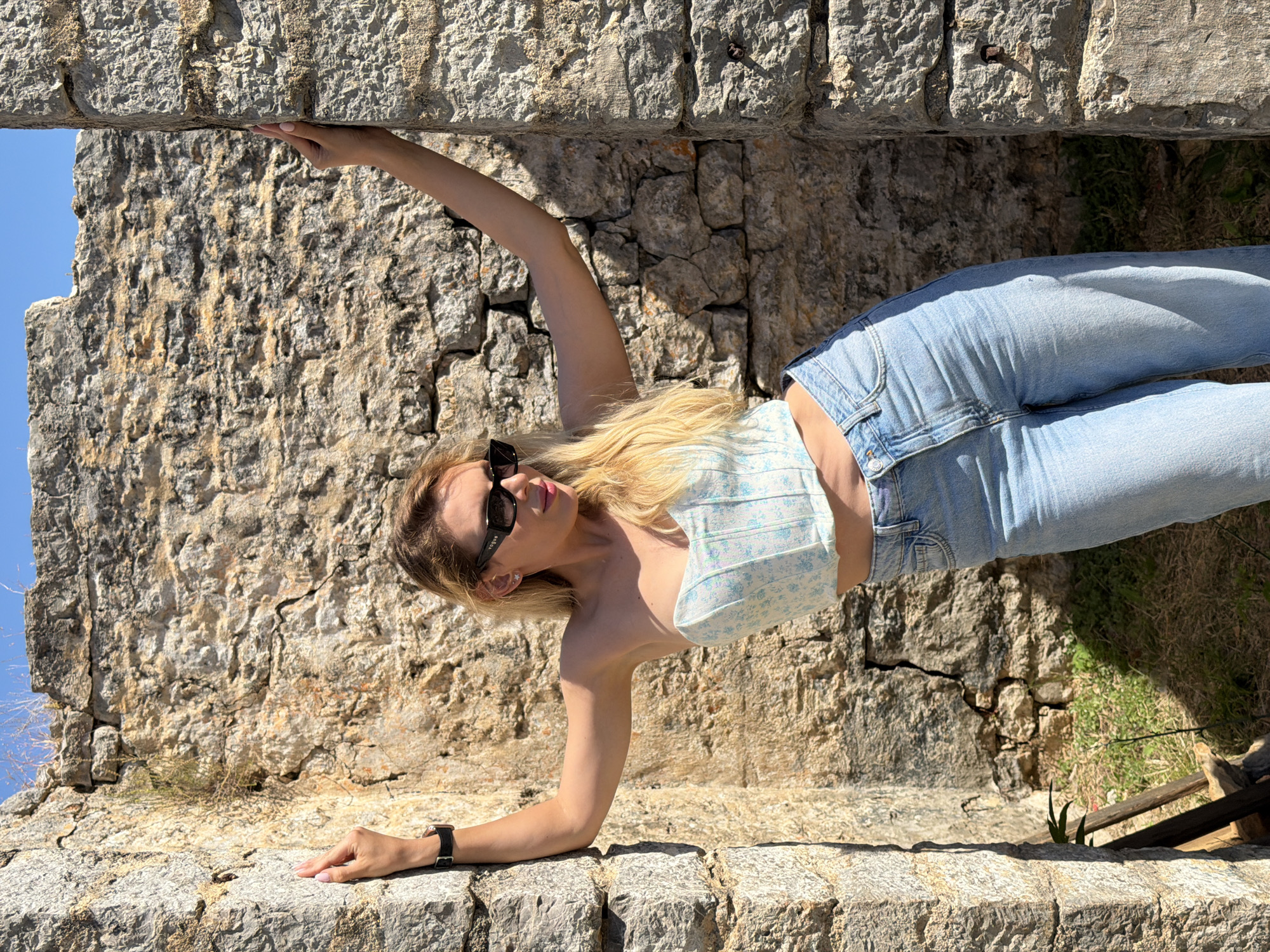 A blonde female is positioned between two weathered stone pillars within an ancient or ruined stone structure, likely in the Old Town of Budva, Montenegro. She is leaning back, with both arms extended upwards and outwards, her hands resting flat against the inner surfaces of the rough-hewn stone columns. Her left wrist displays a black-strapped wristwatch. She wears a light blue and white floral strapless corset crop top, high-waisted light blue denim jeans, rectangular black sunglasses with "Vogue" branding on the right temple, and small stud earrings. Her head is tilted to her right, and she looks in that direction. The stone walls behind her and on either side are irregular, light brown-grey, and textured, showing cracks and natural variations. A clear blue sky is visible above the structure. Sparse green vegetation and dry earth are present on the ground at the base of the walls. Strong sunlight illuminates the scene, creating distinct shadows.