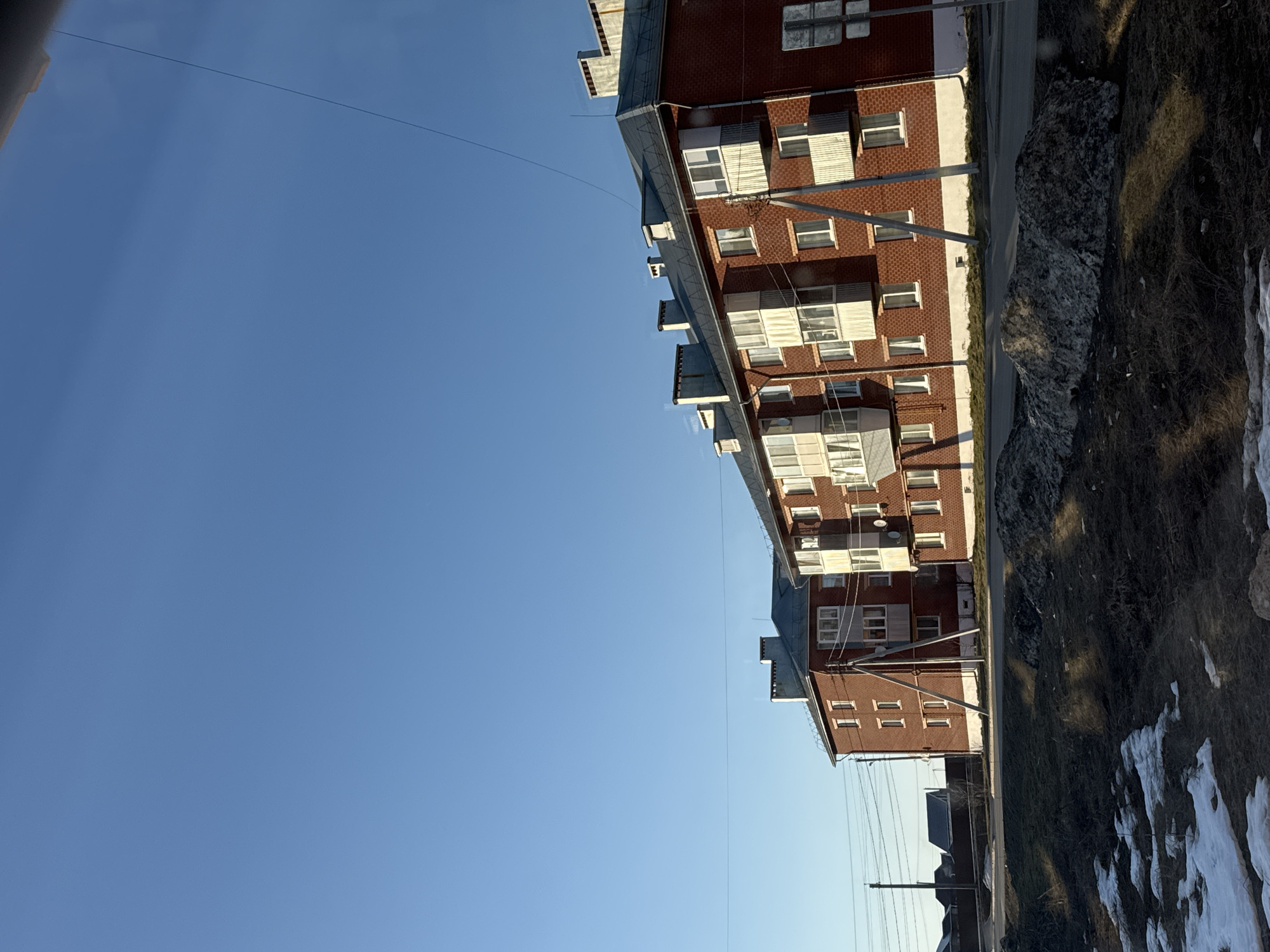 The image shows a clear, blue sky above a row of brick apartment buildings in Chernushka, Russia. The buildings are red brick with white window frames and are likely typical Soviet-era construction. A road runs alongside the buildings, and on the other side of the road is a grassy area with patches of snow, indicating it is likely late winter or early spring. Overhead power lines are visible stretching across the sky, running parallel to the buildings.