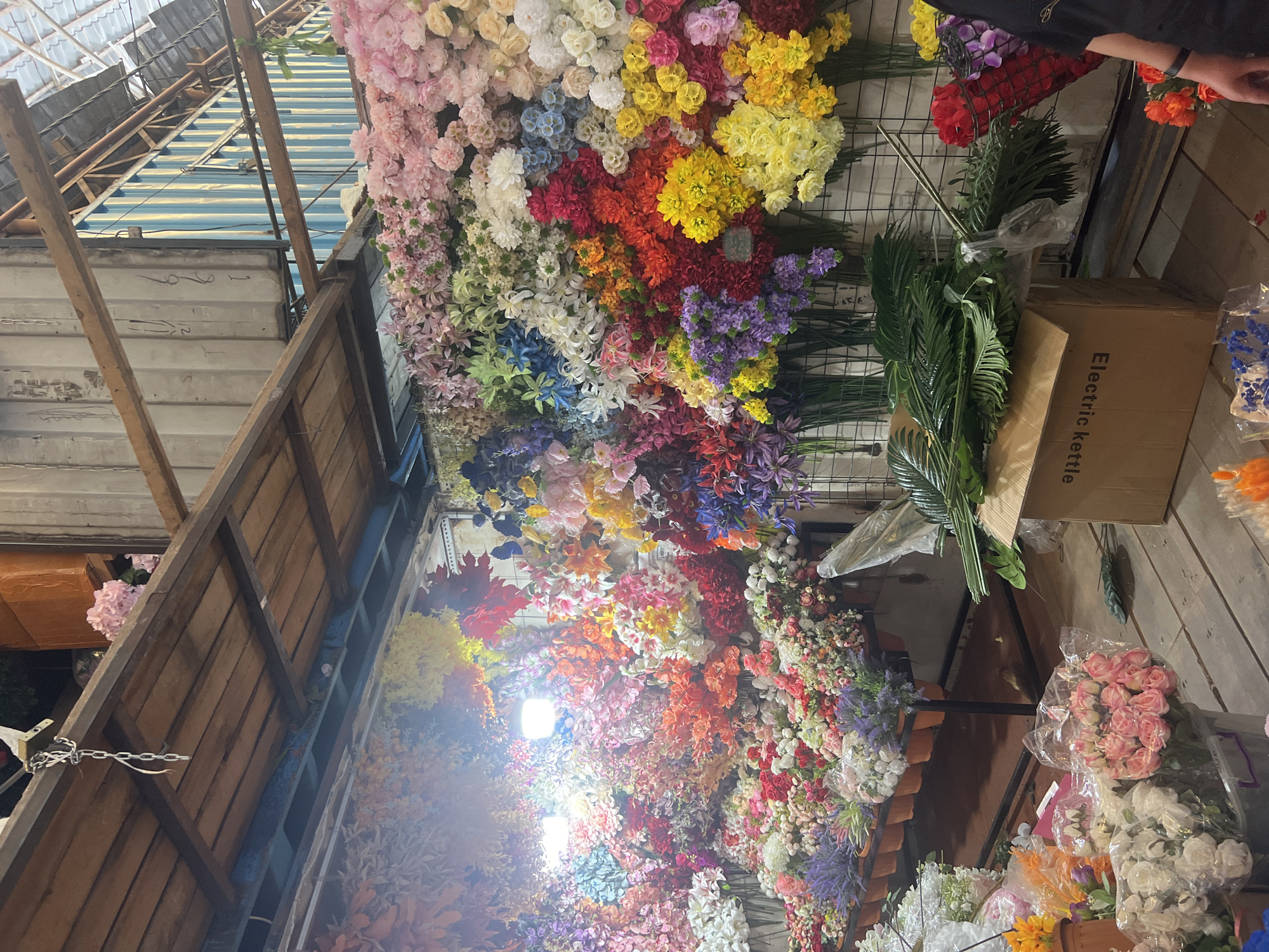 This image depicts a vibrant and abundant display of artificial flowers, likely at a market stall in Bishkek City, Kyrgyzstan. The stall is overflowing with a wide array of colorful flowers, ranging from soft pinks and whites to bright reds, yellows, and blues. They are arranged in a dense and layered fashion, with some hanging from the ceiling and others displayed on shelves and in buckets.
Several large bunches of flowers are visible in the foreground, including pink roses and white roses, packaged in plastic. Behind these, a long display shelf holds potted flowers, with lavender and other smaller blooms. A cardboard box with "Electric Kettle" printed on it sits on the floor, partially covered by large green artificial leaves.
The background reveals the structure of the stall, with wooden beams and a corrugated metal wall. The lighting is a mix of ambient light and overhead artificial lights, creating a warm and inviting atmosphere. No people are clearly visible in the scene, though the presence of the displayed goods suggests a commercial setting. The overall impression is one of a lively and colorful flower market.