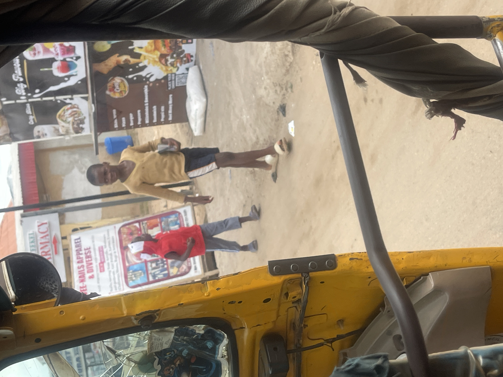 This is a scene on a dirt road in Abuja, Nigeria, as seen from inside a yellow auto-rickshaw. In the foreground, the weathered yellow paint of the auto-rickshaw's interior is visible. A dark gray bar runs across the frame, partially obscuring the view outside.
Outside the auto-rickshaw, two young boys are present. One boy, wearing a red vest and a white hat, stands facing left. He is wearing gray trousers and appears to be looking down. The second boy, slightly taller and wearing a mustard-yellow long-sleeved shirt and dark shorts with a white stripe, is walking towards the right. He holds a dark rectangular object, possibly a book or tablet, in his right hand. He is wearing light-colored sandals.
In the background, there are several signs. One sign reads "PHARMACY" with a red arrow pointing to the right. Below it is a larger sign advertising "FEE-NAILS APPAREL & DIVERSE". To the right, a food stall is visible with menus and images of food items like burgers, fries, and drinks. A blue trash bin is next to the stall. The ground is covered in light brown dirt, with some scattered paper debris. The sky is overcast.
