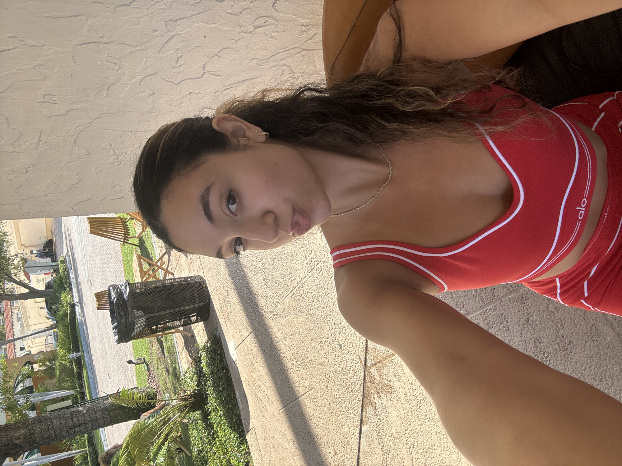 In Boca Raton, Florida, a young woman with long, wavy brown hair and fair skin takes a selfie outdoors. She is wearing a red athletic bra with white trim and a white logo that reads "alo." Her lips are puckered, and she is wearing a delicate gold necklace and small hoop earrings. Behind her, a textured cream-colored wall serves as a backdrop. To her left, the scene transitions to an outdoor setting with a paved walkway, green foliage, a trash can, and wooden chairs and tables. The sun casts distinct shadows on the walkway. In the background, a street is visible with trees, buildings with red tiled roofs, and vehicles.
