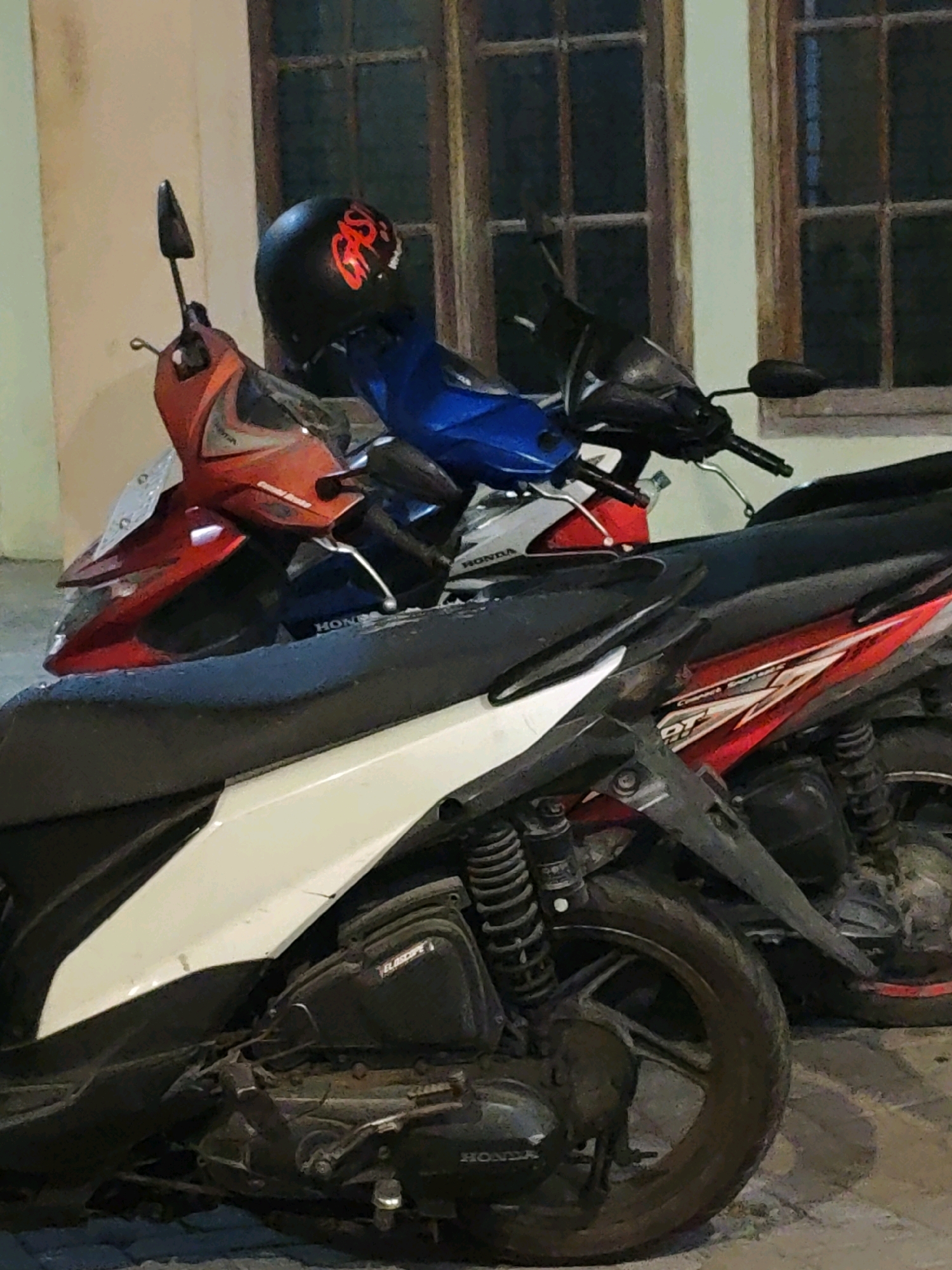 The image captures a close-up view of several parked scooters in what appears to be a residential area or a small business in Ngasem, Indonesia. The scooters are positioned tightly together, with their handlebars interlocked or overlapping, suggesting a communal parking arrangement.
In the foreground, a dark-colored scooter with a white accent panel dominates the lower half of the frame. Its engine and rear wheel are visible, showing signs of wear and tear. The seat is a matte black.
Behind this scooter, partially obscured, are two other scooters. One is predominantly blue, with a black handlebar and mirror. A black helmet with red lettering is perched on its seat. The other scooter visible is bright red and orange, with black trim. The front of this scooter, including its headlight and fairing, is visible.
Further in the background, another scooter, this one predominantly red with white and black accents, is partially visible, its rear section and wheel showing. The phrase "SPORT" is visible on its side panel.
The scooters are parked on a paved surface, possibly a sidewalk or a small alleyway. Behind them, a building facade with wooden-framed windows is visible. The lighting suggests it is either early morning or late evening, with artificial light illuminating the scene. The overall impression is one of a typical everyday scene in a bustling Indonesian town.