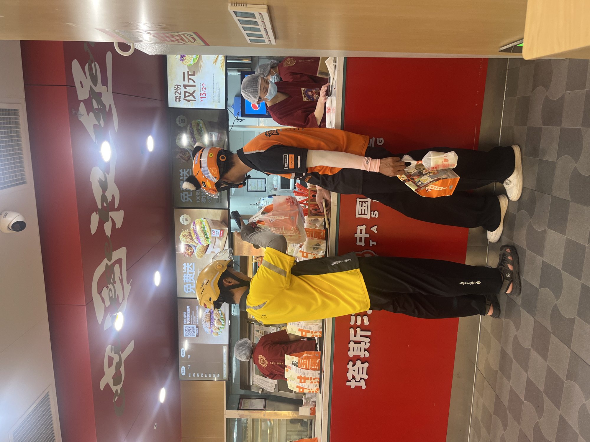 The image captures an indoor scene at a "TASTING" (塔斯汀) fast-food restaurant in Yuzhong District, China. Two male food delivery drivers are positioned at a red service counter. The driver on the left wears a yellow safety helmet, a yellow and black short-sleeved shirt, black pants, and grey sandals, holding a clear plastic bag containing multiple items. The driver on the right wears an orange safety helmet with black cat-ear accessories, an orange and black short-sleeved shirt, black pants, and white sneakers, holding a small brown takeout bag and a plastic cup in a clear bag. Behind the counter, two female restaurant staff members are visible, both wearing maroon short-sleeved uniforms and white hairnets. One staff member, on the left, is partially obscured with her back mostly to the camera, standing behind several brown paper takeout bags. The other staff member, on the right, wears a light blue surgical mask and is looking down, engaged with a counter surface.
Above the counter, multiple illuminated digital displays present food advertisements and promotional offers, including "免费送" (Free Delivery/Gift) and "第2份 仅1元" (Second item only 1 Yuan), along with order status information like "备餐中" (Preparing) and "请取餐" (Please pick up order) with associated numbers. The red counter features the restaurant's name "塔斯汀" in white Chinese characters. The ceiling is red with white, abstract decorative elements, recessed lighting fixtures, a security camera, and an air vent. The floor is tiled with a patterned design of grey and darker grey squares and curved shapes. A light wooden paneling with a white rectangular wall unit is visible on the right side of the frame.