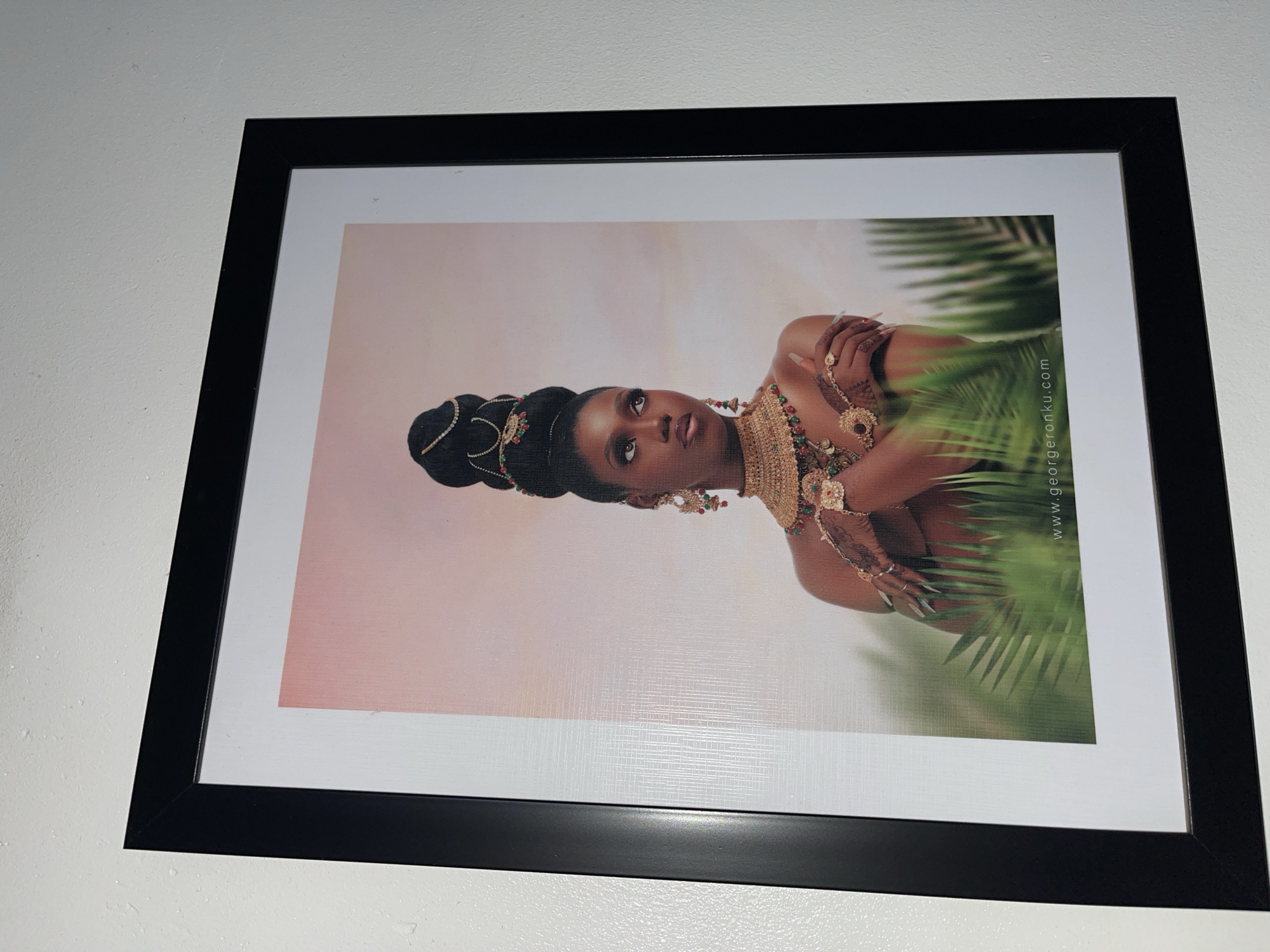 A framed portrait of a woman hangs on a plain white wall, captured from a slightly angled, upward perspective. The frame is thick and black.
The framed artwork depicts a young Black woman adorned in elaborate, cultural attire. Her hair is styled in a tall, intricately braided or wrapped updo, accented with gold bands and small red and green beads. She wears a striking golden necklace with matching red and green gemstone details, large ornate earrings, and numerous gold rings and a delicate hand chain (hathphool) on her hands. Her long fingernails are painted a light color. She is shown from the chest up, with her head slightly tilted and her gaze directed towards the viewer, wearing a serene and regal expression.
The background of the portrait features a soft, gradient sky in shades of light pink and peach, suggesting either dawn or dusk. On the right side of the image, lush green palm-like fronds are visible, adding a tropical or natural element to the scene.
A visible web address, "www.georgeronku.com", is printed vertically along the right edge of the portrait, likely crediting the artist or photographer. The print itself appears to have a subtle woven or canvas-like texture. The overall image captures this piece of art displayed in what appears to be an indoor setting.