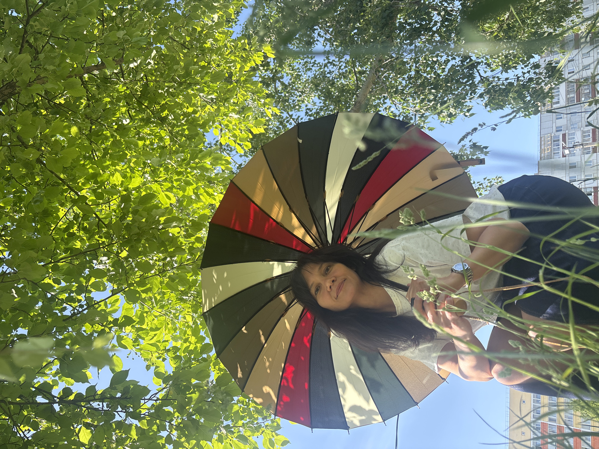 A low-angle shot depicts a woman holding an open, multicolored umbrella, with a tree canopy and a building visible in the background. The woman has dark hair and is wearing a light-colored shirt and dark bottoms. The umbrella has alternating stripes of brown, tan, green, red, and gray colors. The tree leaves are vibrant green, and the sky is a clear blue. Tall blades of grass frame the lower portion of the image. The building in the background appears to have multiple stories and numerous windows. The location is Chaikovsky, Russia.