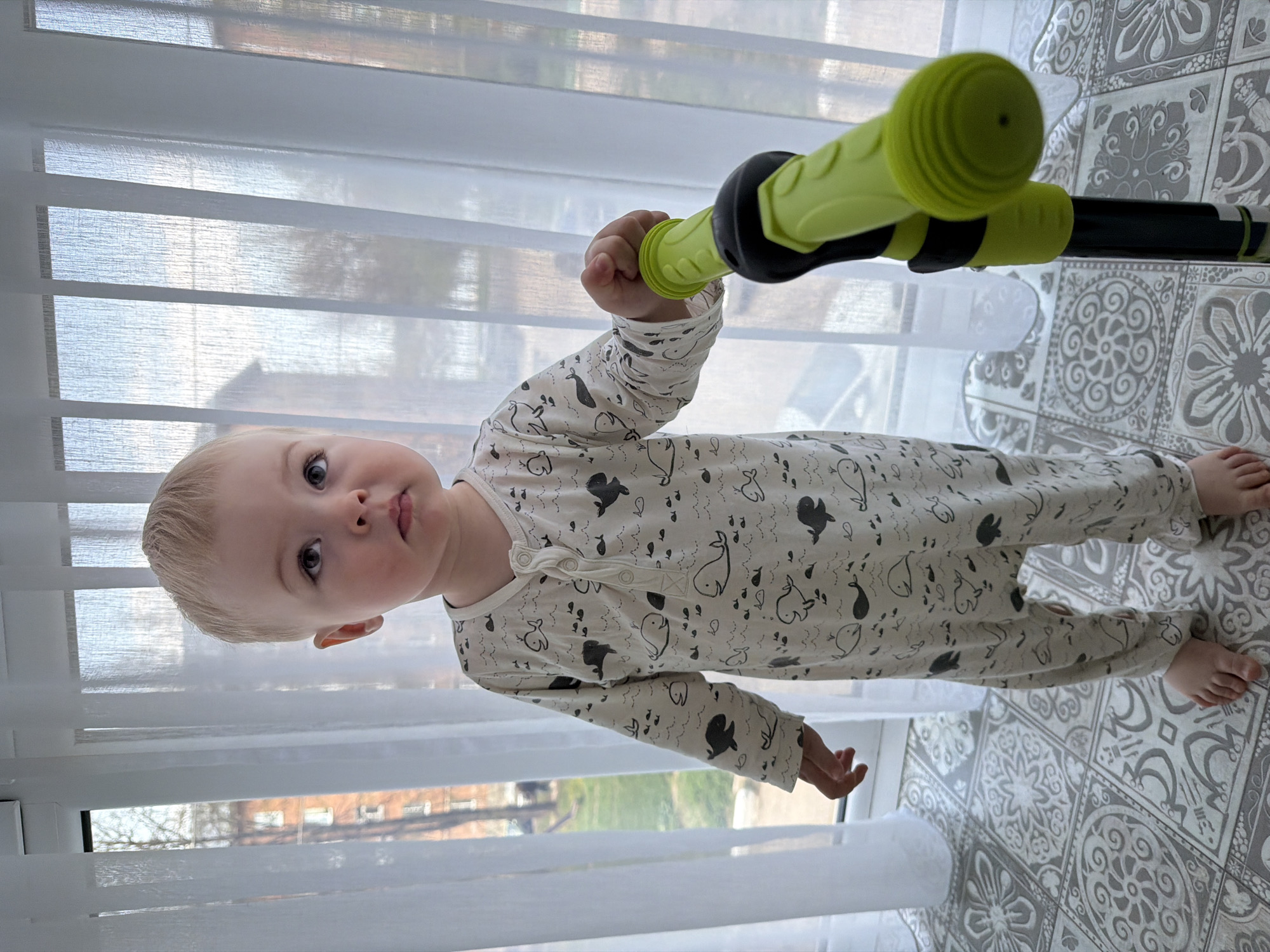 A fair-skinned, blond toddler wearing pajamas with a whale pattern stands indoors on a gray and white tile floor. The child holds the neon green handle of a black and green scooter, looking directly at the camera. A bright window with sheer white blinds is visible behind the child, with a glimpse of the exterior showing trees and a building. The location is unknown, and the description cannot be narrowed to the city of Votkinsk, Russia.