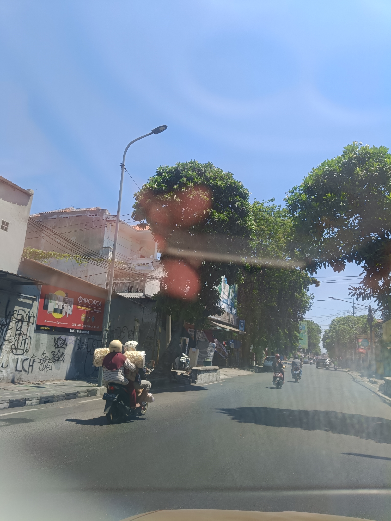 The image shows a bright, sunny day on a street in Sidoarjo, Indonesia. The viewpoint appears to be from a vehicle moving along the road. On the right side of the road, there are several trees with lush green foliage, with a bright blue sky above. Buildings and storefronts line the left side of the road, some adorned with graffiti. A prominent red and white sign for "IMPORTA BUKIT FURNITURE" is visible on the left.
In the foreground, two individuals are riding a scooter. The person in the back is wearing a maroon shirt and a yellow helmet, while the person in the front is wearing a white helmet and is mostly obscured. They are carrying a large amount of what appears to be packaged snacks or food items, wrapped in clear plastic.
Further down the road, several other scooters and some cars can be seen, indicating moderate traffic. The road surface is asphalt, with a white line marking the lane. The overall scene depicts a typical street view in a bustling Indonesian city on a clear day.