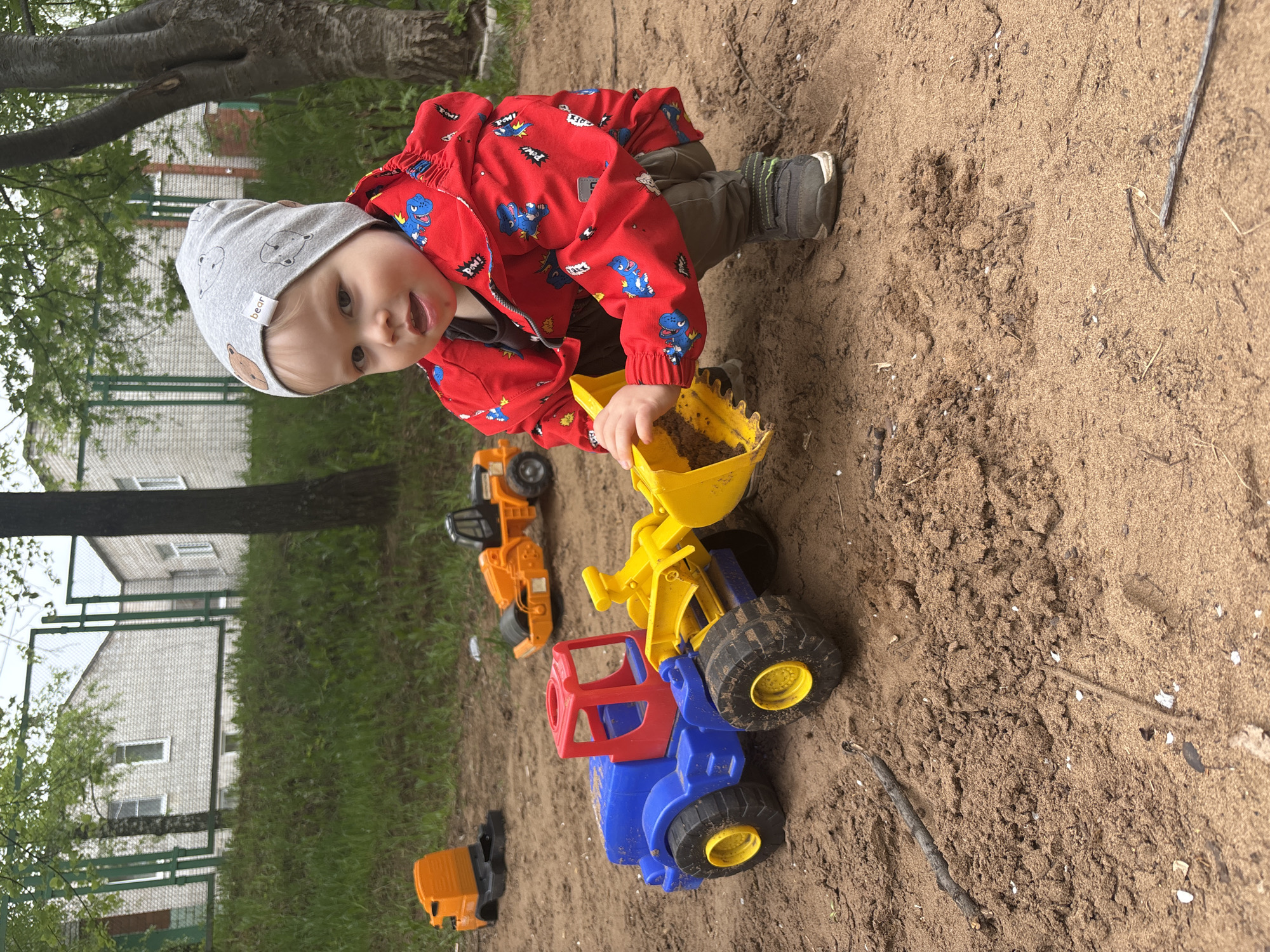 A toddler in a red dinosaur-patterned jacket and gray bear-patterned beanie is playing in a sandbox, filling a yellow toy scoop with sand. He is leaning over a large, blue and yellow toy truck. Several other orange toy construction vehicles are scattered nearby in the sand. The scene appears to be outdoors, possibly a playground in Izhevsk, Russia, based on the visible architectural style of the building and fence in the background. The environment includes grass and a tree.