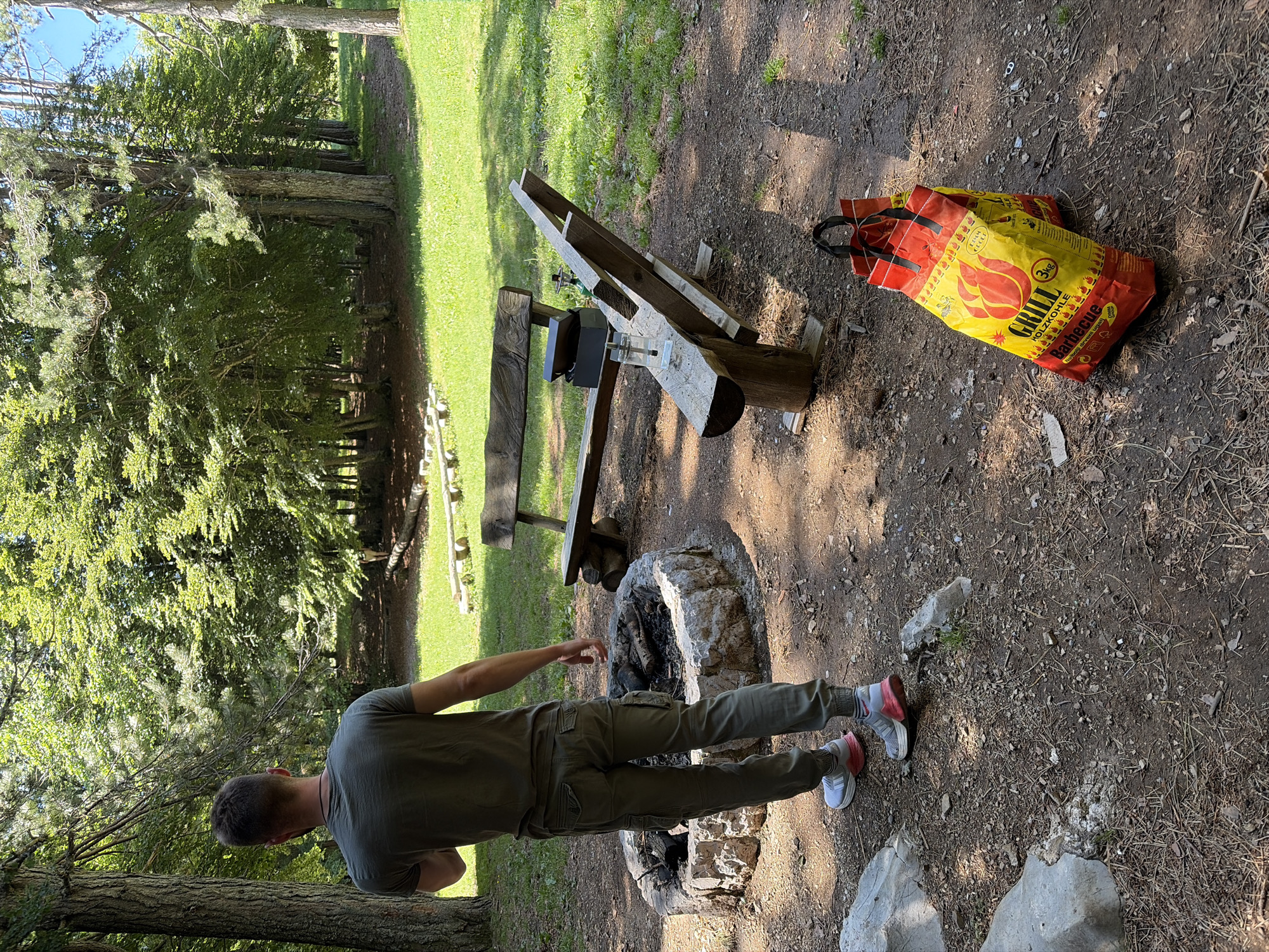 The image captures an outdoor recreational area in Dolovi, Montenegro, under bright daylight. A male individual, seen from behind, is positioned slightly left of center, facing into a dense forest. He wears a dark olive-green short-sleeved t-shirt, olive-green cargo pants with elastic cuffs, and grey and red athletic shoes. In the lower-left, a circular fire pit constructed from rough-hewn stones contains partially burnt wood. To the right of the fire pit, two rustic wooden benches are present. A clear plastic water bottle or glass with a metal straw, a black rectangular box, and a small green cylindrical object are placed on the longer wooden bench. Two yellow and red bags of "GRILL HOLZKOHLE Barbecue 3KG" charcoal lie on the dirt ground in the lower-right quadrant. The immediate ground cover is dry dirt, pine needles, and small stones, transitioning to green grass and a dense background of mixed deciduous and coniferous trees. Distinct shadows are cast from the benches and the charcoal bags. In the far background, a faint dirt path leads into the forest, with a barely discernible figure further down the path.