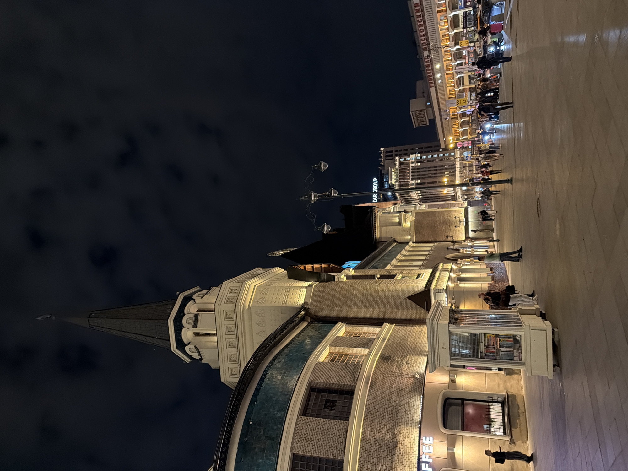 The image depicts a nighttime street scene in Moscow, Russia, specifically at the Moskovsky railway station. In the foreground, a street vendor's kiosk, resembling a small white booth with glass panels displaying books, stands on a paved plaza. Several people are visible: one individual stands near the kiosk, another walks away from it, and a group is gathered further down the plaza. To the left, a building with a prominent arched facade and windows is lit up, with signage for "COFFEE." Above this, the ornate spire of the station building pierces the dark, cloudy night sky. To the right, more buildings are illuminated, including a tall modern structure with many windows, and across the plaza, the facade of the railway station is brightly lit with many windows and signs. Cars are parked and moving in the background, suggesting a busy urban environment. The overall atmosphere is that of a well-lit, active public space at night.