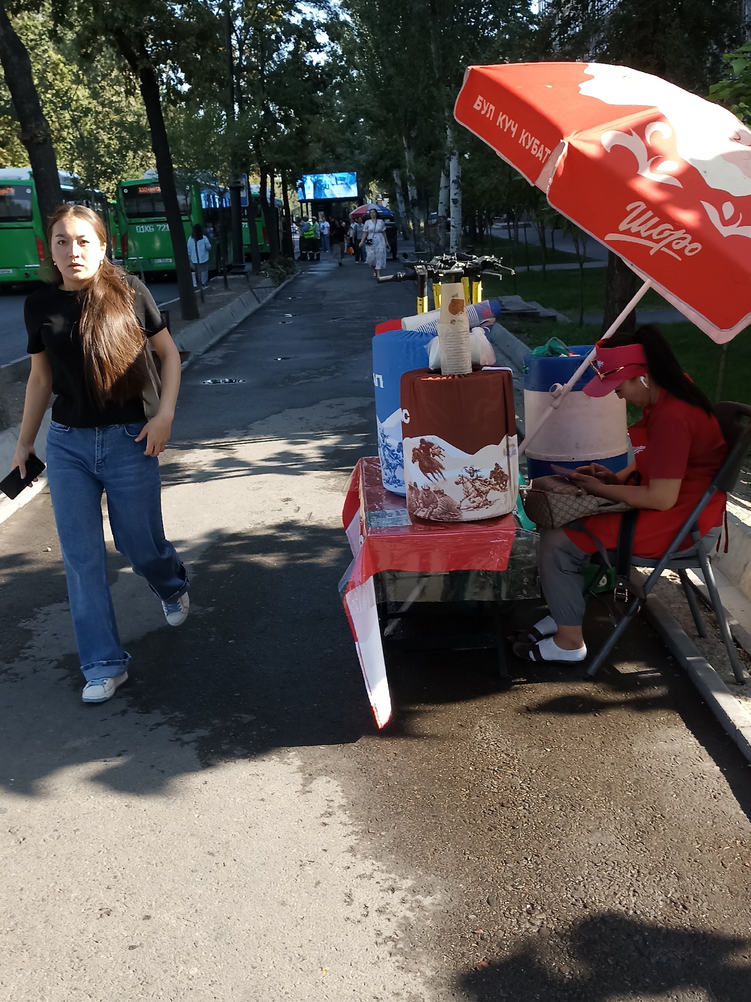 A woman in a black t-shirt and blue jeans walks down a sidewalk in Bishkek, Kyrgyzstan, holding a black smartphone. To her right, a vendor sits on a chair behind a table displaying various items, including large blue and white containers, a red umbrella with Cyrillic writing, and a brown box with an illustration of cowboys. The vendor wears a red shirt and a pink visor, looking down at her own phone. Several green buses are visible in the background, parked along the street. Trees line the sidewalk, and the ground is paved with asphalt and concrete. The scene appears to be a street-side market or vendor stall on a sunny day.