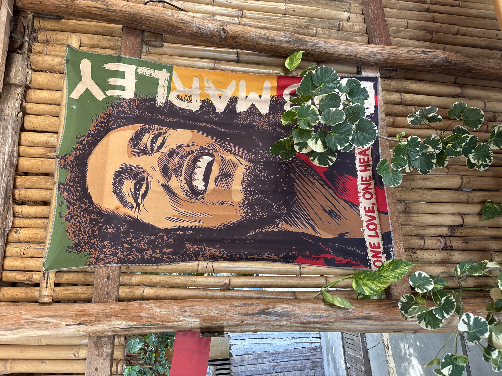 A Bob Marley poster is mounted on a bamboo structure in Baan Bo Phut, Thailand. The poster is vibrant with red, yellow, and green hues, displaying a portrait of Bob Marley and the text "ONE LOVE. ONE HEART." A leafy plant drapes across the right side of the poster and onto the bamboo. The bamboo forms part of a hut-like structure. The scene is partly shaded.