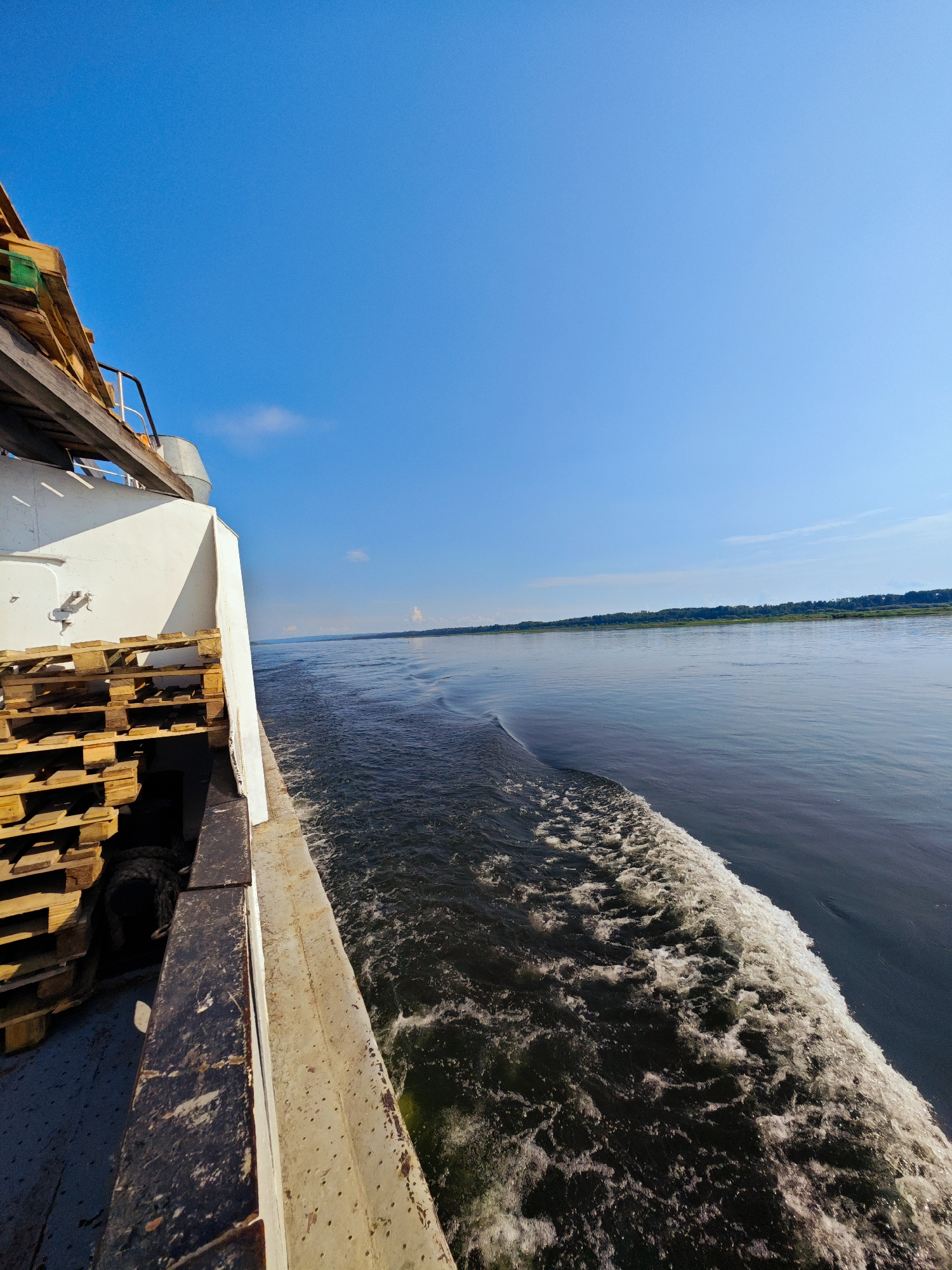 The image shows a wide, calm river on a sunny day. The camera is positioned on the deck of a boat, looking towards the water and the distant shore. On the left side of the frame, the side of the boat is visible, with a stack of wooden pallets. The boat is moving, creating a wake in the water. The sky is a clear blue with a few wispy clouds. The distant shore is lined with trees. The scene appears to be in Novonazimovo, Russia.