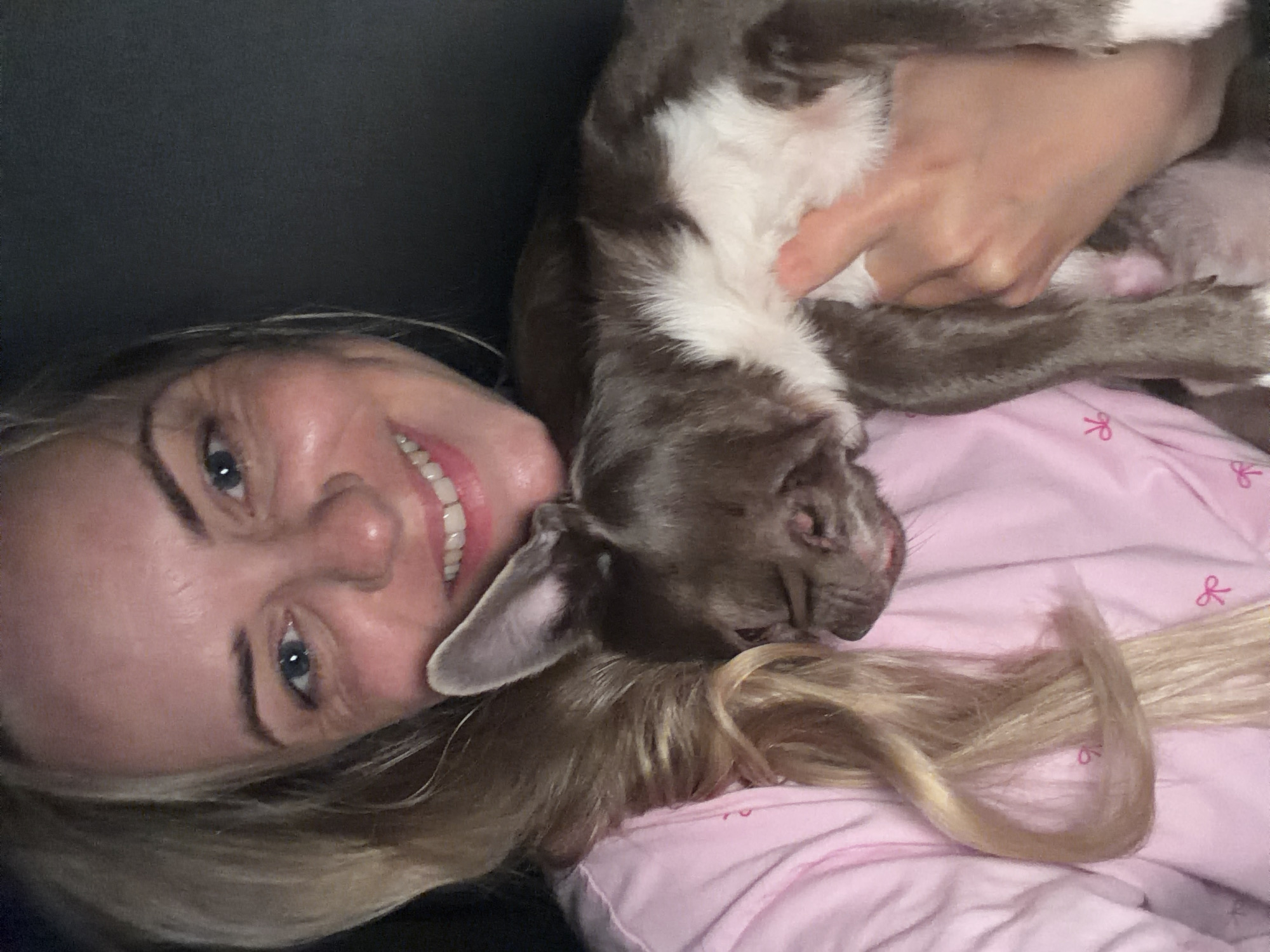 A medium shot taken from a high angle shows a Caucasian woman with blonde hair and blue eyes holding a small dog. The woman is smiling and looking at the camera, while the dog is resting in her arms, with its eyes closed. The woman is wearing a pink pajama top with a bow pattern. The background is dark and out of focus, making it difficult to determine the location. However, if this photo was taken in Votkinsk, Russia, it is likely an indoor setting, such as a bedroom or living room. The lighting suggests it may be nighttime or a dimly lit room. The overall mood of the image is warm and affectionate, conveying a sense of comfort and companionship between the woman and her pet.