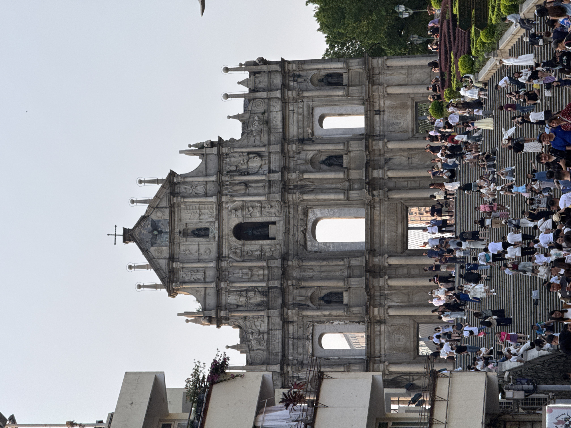 The image features the Ruins of St. Paul's in Macau, China, with a large crowd of people on the steps leading up to the facade. The ruins consist of a stone facade with intricate carvings and architectural details, including statues, columns, and arched windows. The crowd comprises people of various ages and ethnicities, dressed in casual clothing. Some are taking pictures, while others are simply walking up or down the steps. The environment includes trees and landscaping on the sides of the steps. The sky is bright and clear. Modern buildings are seen at the base of the ruins.