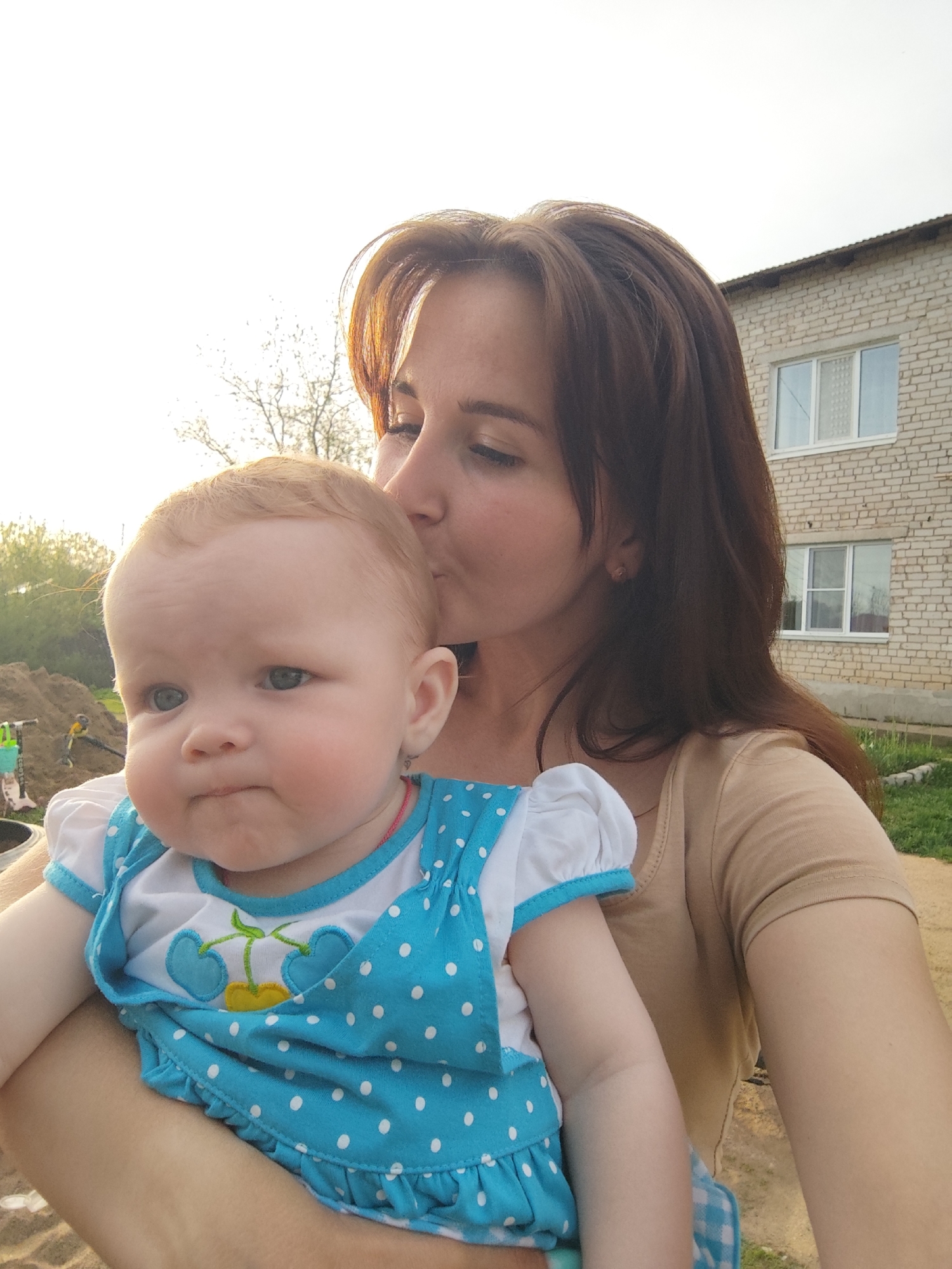 A woman is holding a baby, appearing to kiss its head. The baby has fair skin, light hair, and is wearing a blue polka-dot dress. The woman has brown hair, fair skin, and is wearing a light brown shirt. Behind them, there is a two-story brick building with two visible windows. To the left, there is a pile of dirt and some shrubbery. The picture seems to be taken outdoors during daytime in Фоки, Russia.