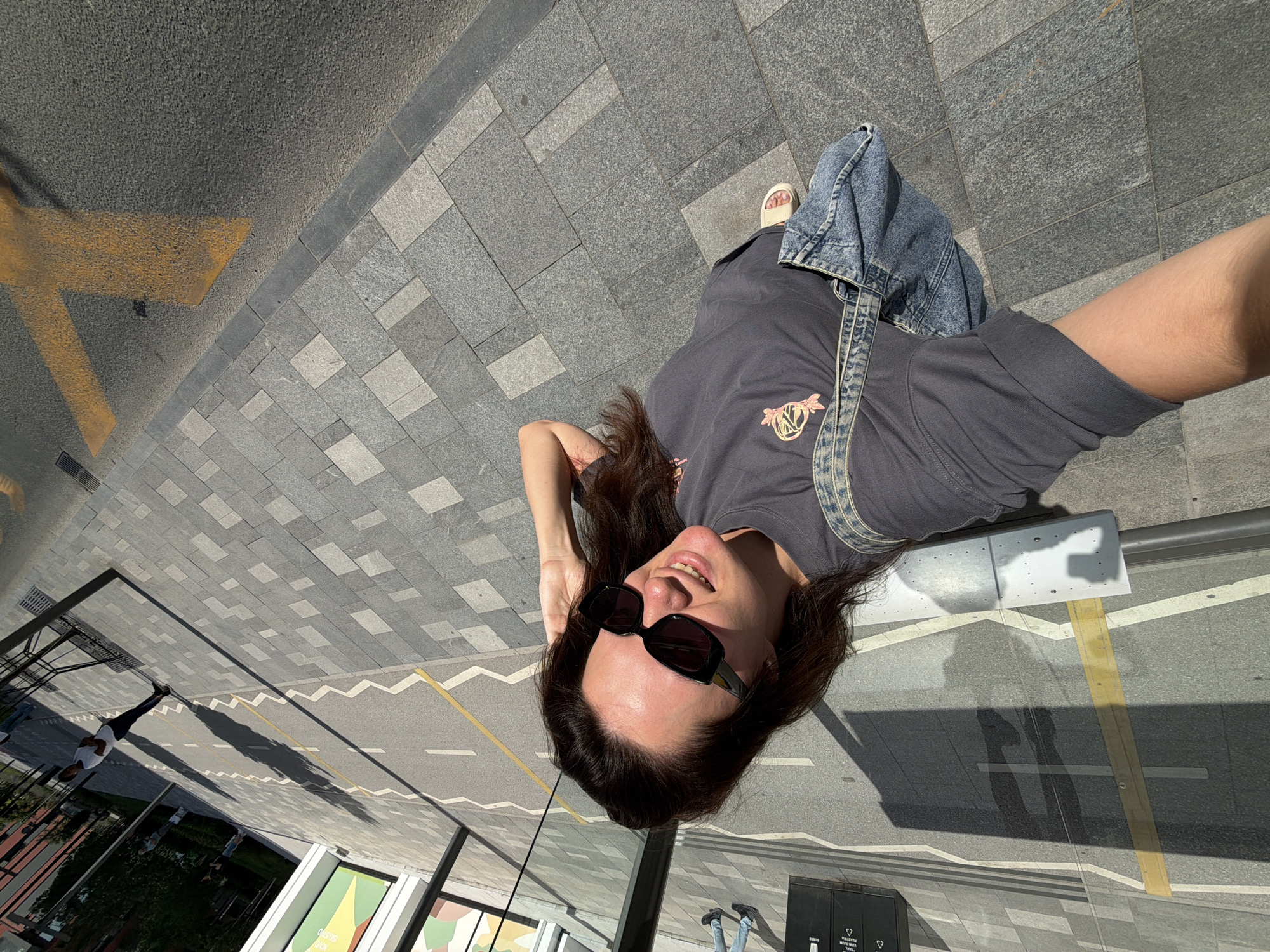 In an outdoor urban setting in Belgrade, Serbia, a woman with long brown hair and sunglasses is taking a selfie. She is wearing a dark gray t-shirt and a denim shoulder bag. She stands on a paved area with a patterned, grey stone surface. In the background, a street with yellow markings and a sidewalk with a zigzag pattern is visible, along with reflections and shadows from buildings and people. A black waste bin is also present.