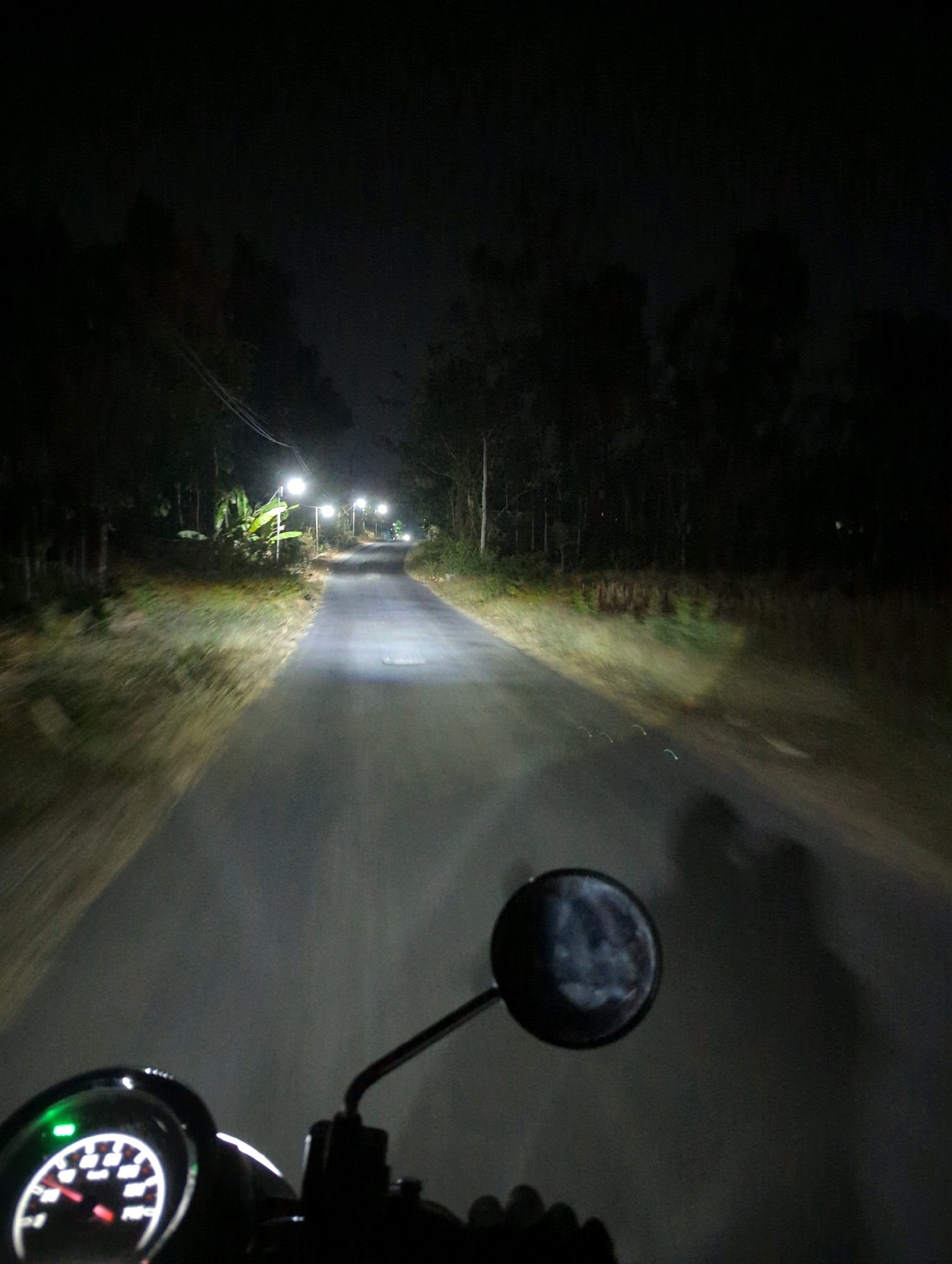 An over-the-shoulder perspective shot captures a dark, paved road at night, illuminated by streetlights and the headlight of the viewer's vehicle, likely a motorcycle given the presence of a handlebar and a speedometer in the foreground. The road curves gently, disappearing into the darkness ahead. The sides of the road are lined with dense, dark trees, and in the mid-ground to the left, there is a cluster of banana trees under a streetlight. Several streetlights are visible along the road, casting pools of light. The speedometer shows a red needle pointing to approximately 20. The overall impression is of a rural road at night, possibly in or near Senggrong, Indonesia, judging by the vegetation and infrastructure.