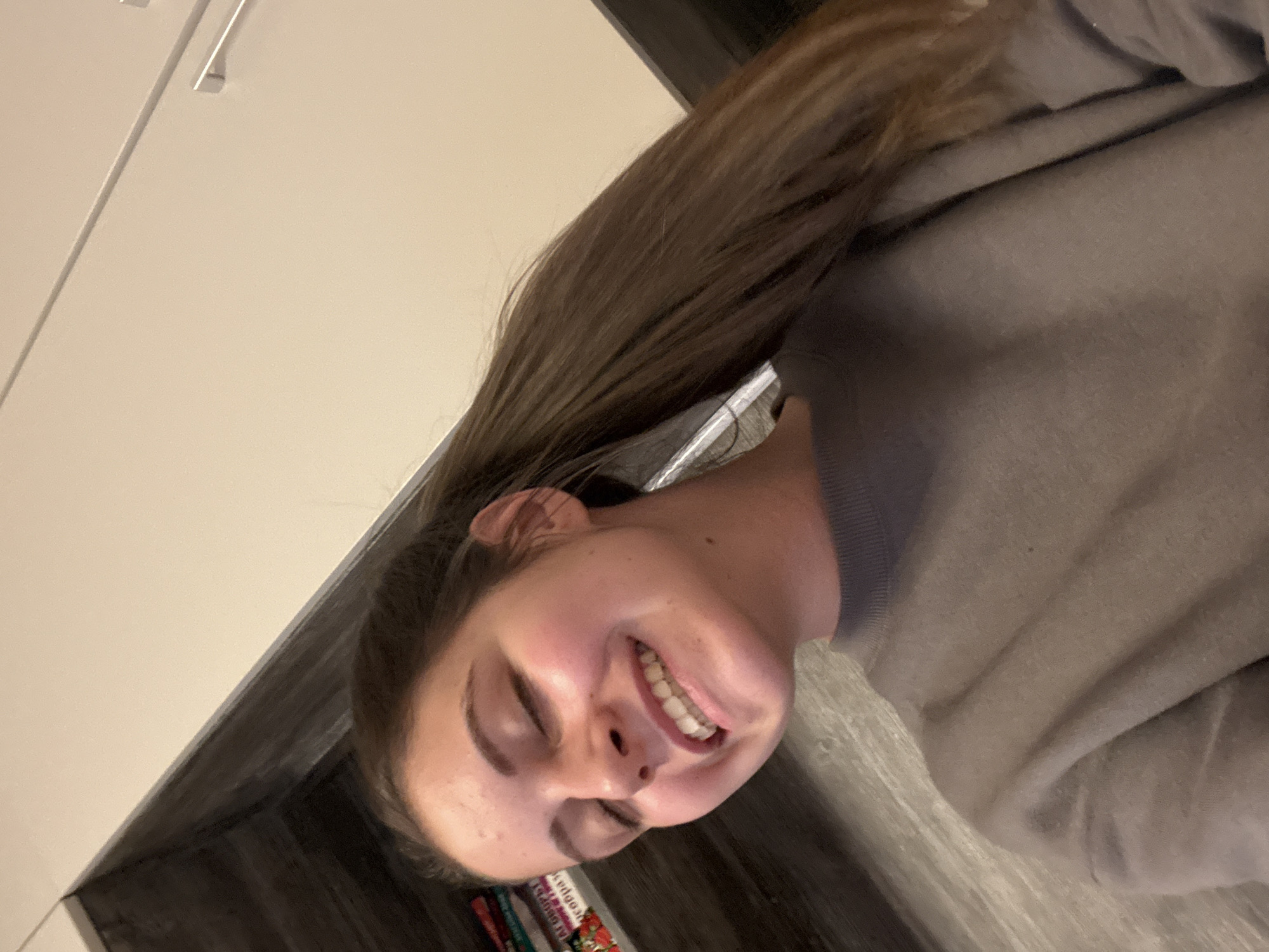 A rotated close-up portrait captures a young person, likely female, in a moment of candid joy or laughter. Her head is tilted, and her eyes are closed as she displays a wide, natural smile, revealing her upper teeth. She has long, straight brown hair that cascades over her shoulder and back, and she is wearing a plain grey or taupe long-sleeved top.
The setting is indoors, with light-colored, possibly white, modern cabinet doors or walls visible in the background to her upper left, featuring subtle linear details and what appear to be sleek silver handles. Below and to the right, darker wooden or tiled surfaces are present. In the lower left foreground, below the person's head, a stack of colorful objects, potentially books or packages, can be seen. Partially visible text on these items includes fragments such as "EDGE" and "AFG."
The lighting indicates an indoor environment, without specific cues for the time of day. The photograph conveys a feeling of happiness and amusement. The location context provided is Bulgarovo, Bulgaria.
