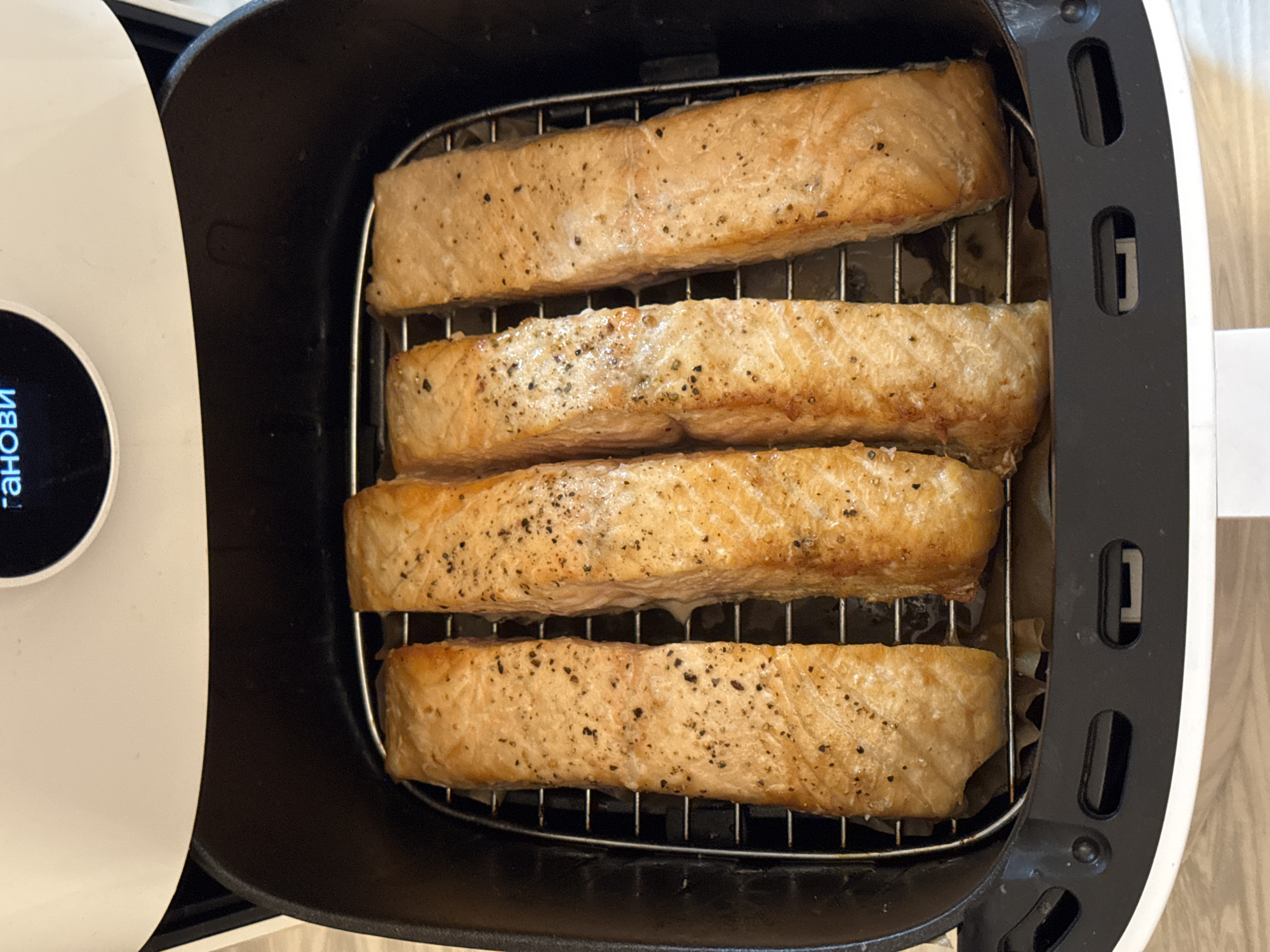 An overhead shot reveals four salmon fillets in an air fryer. The fillets, seasoned with black pepper, rest on a wire rack inside the black basket of the fryer. The fryer's white control panel is visible on the left. The scene is lit with natural light, casting soft shadows within the fryer's interior. The location, Chaikovsky, Russia, is not discernible from the image.