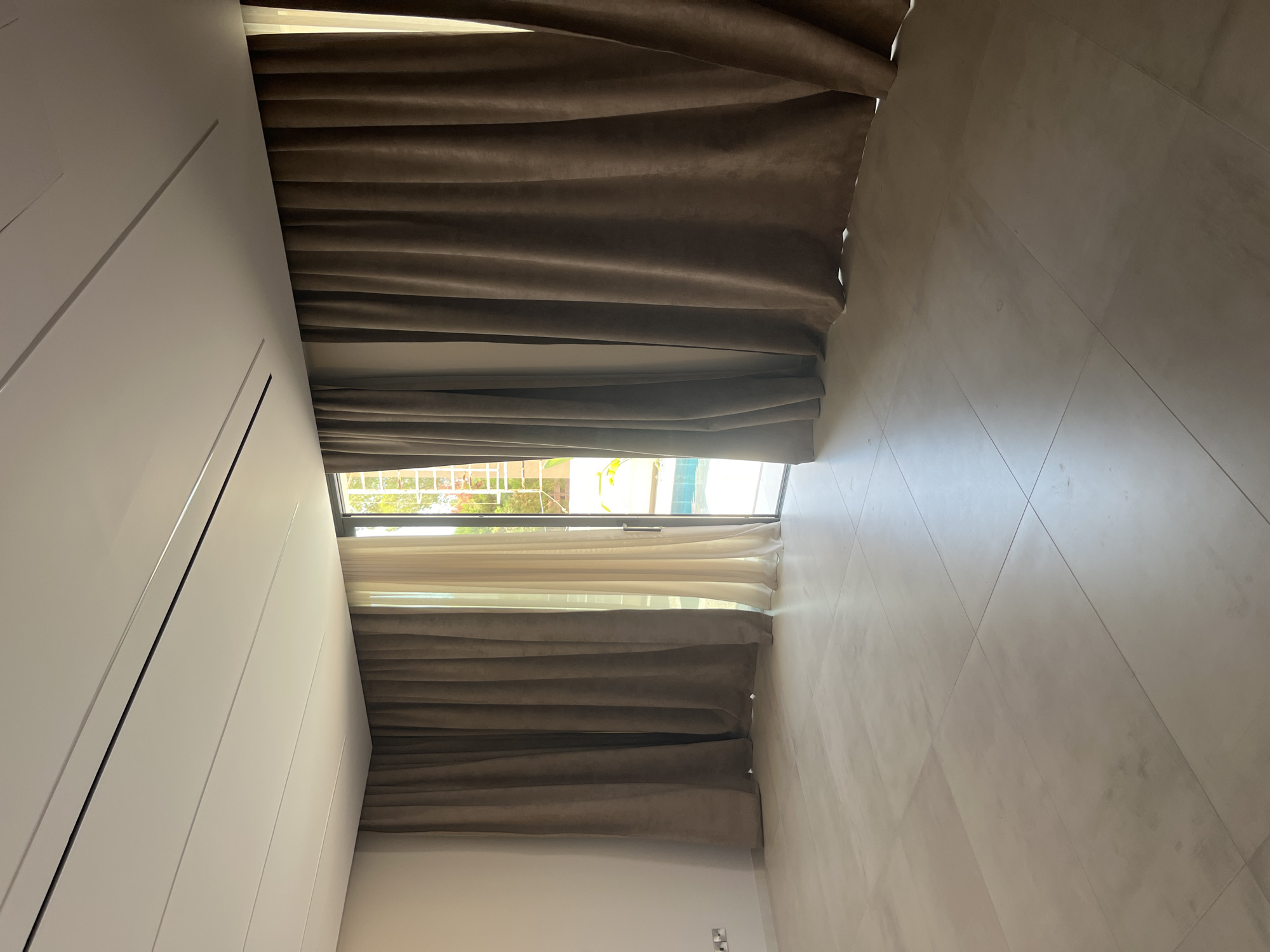 The image depicts an interior view of a room with large sliding glass doors leading to an outdoor pool area. The room has light-colored tiled flooring and cream-colored walls. Two sets of curtains are visible: dark brown, heavy velvet curtains on the right and in the left foreground, and lighter, sheer curtains partially visible on the left. The ceiling is white with subtle linear detailing and integrated lighting. Through the glass doors, a swimming pool with blue water and some surrounding greenery are discernible. The scene is clean and appears to be a residential property in Moni Community, Cyprus, likely a living area or bedroom with a view of the pool. There are no people or animals present in the image.