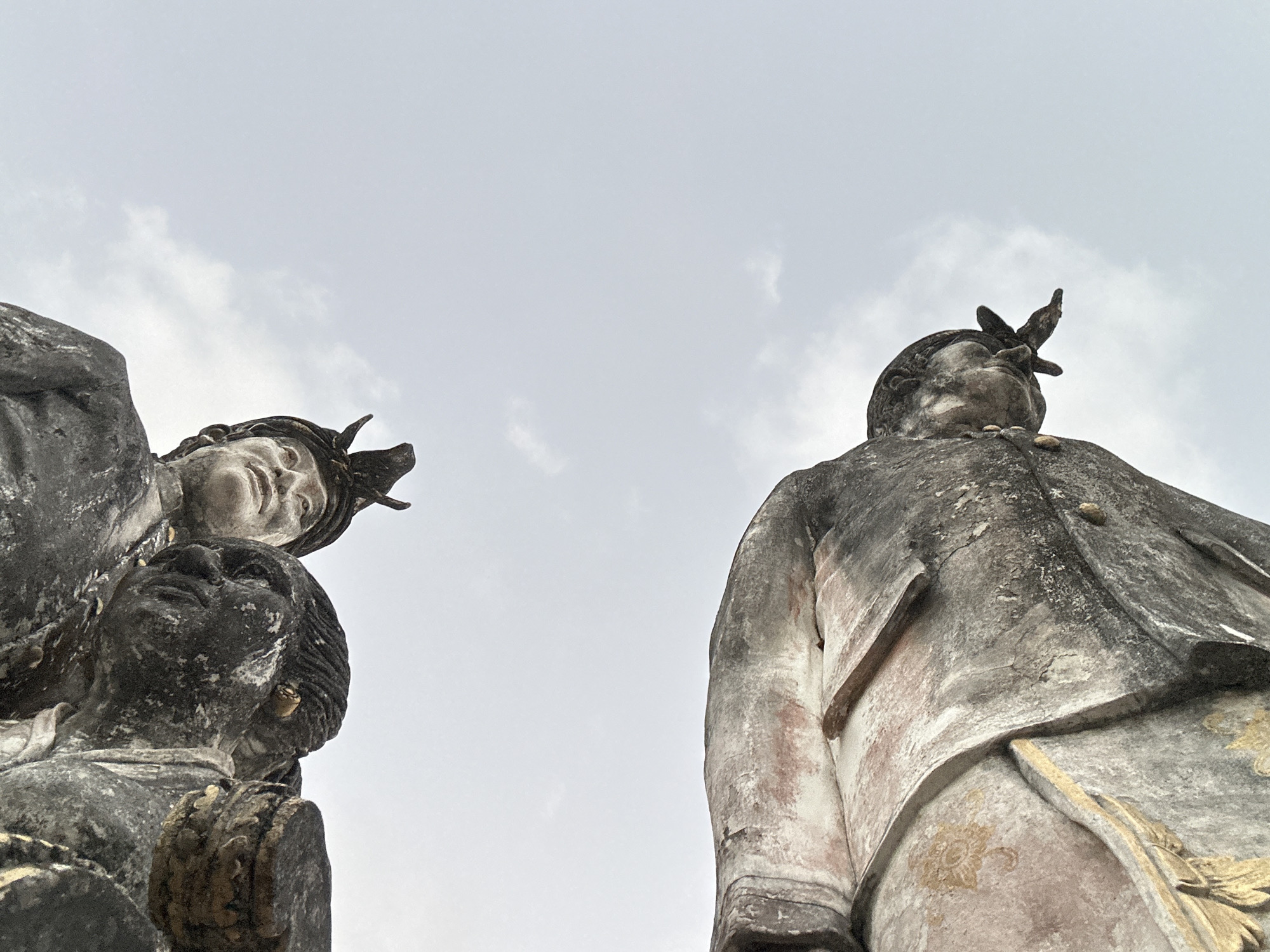 This image shows a low-angle, upside-down shot of two weathered stone statues against a pale, cloudy sky. The statue in the foreground appears to be a reclining figure, its stone body weathered and showing hints of pink and gold decorative elements. The head of this figure is visible, with details of a sculpted face. Above and to the right, another statue is partially visible, also upside-down, showing a carved head with discernible facial features and a headdress. The overall impression is of ancient, possibly religious, figures, weathered by time and elements. Based on the context of Kuta Selatan, Indonesia, these statues are likely part of a temple or significant cultural site, possibly depicting deities or historical figures.