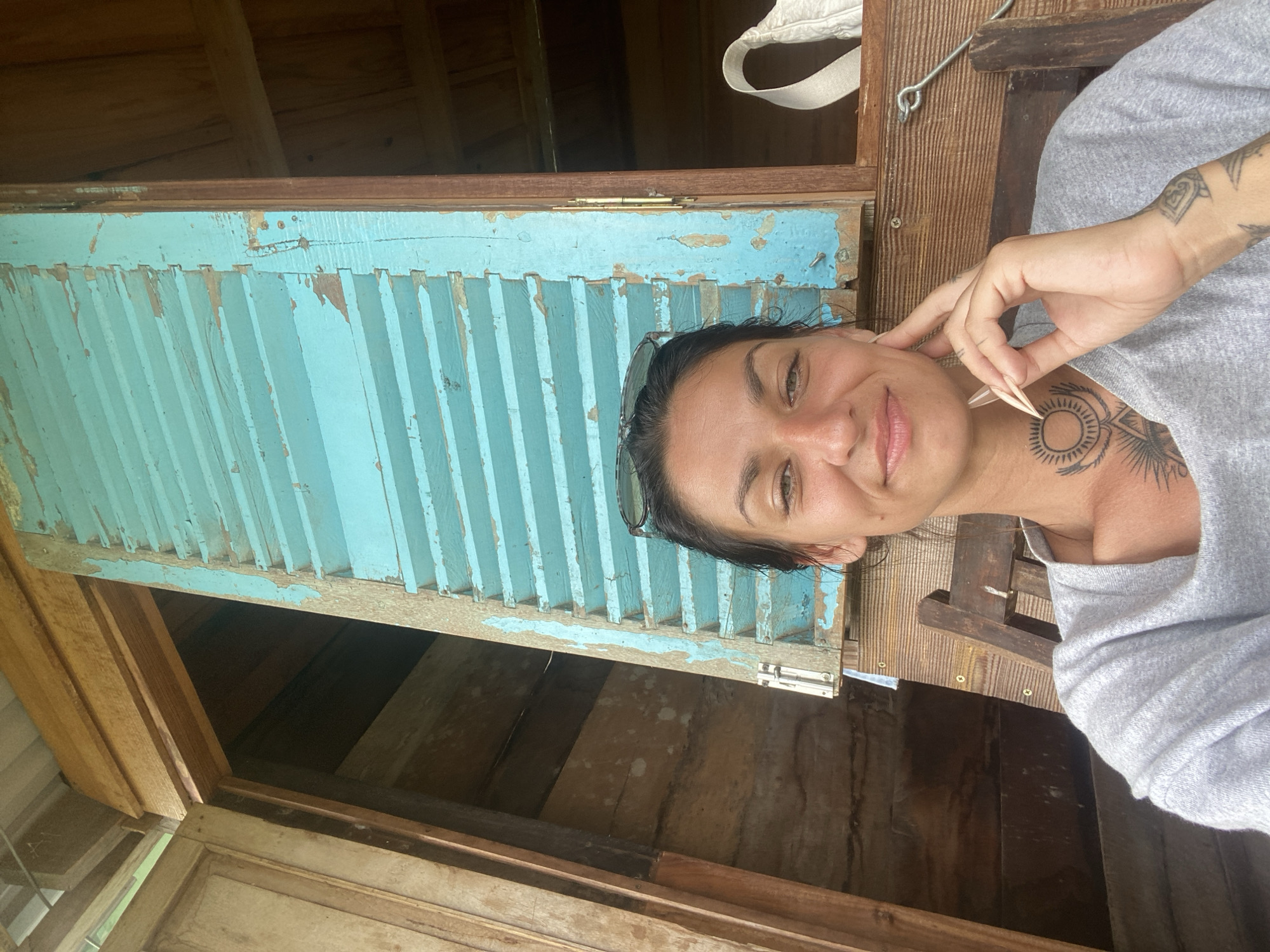 A young woman with dark hair and a tattoo on her neck is sitting on a wooden chair in front of a blue shuttered window. She is smiling and looking at the camera, with her hand resting on her chin. The background is a wooden structure with a thatched roof, possibly a house or a cafe. The overall impression is casual and relaxed, possibly a vacation photo.