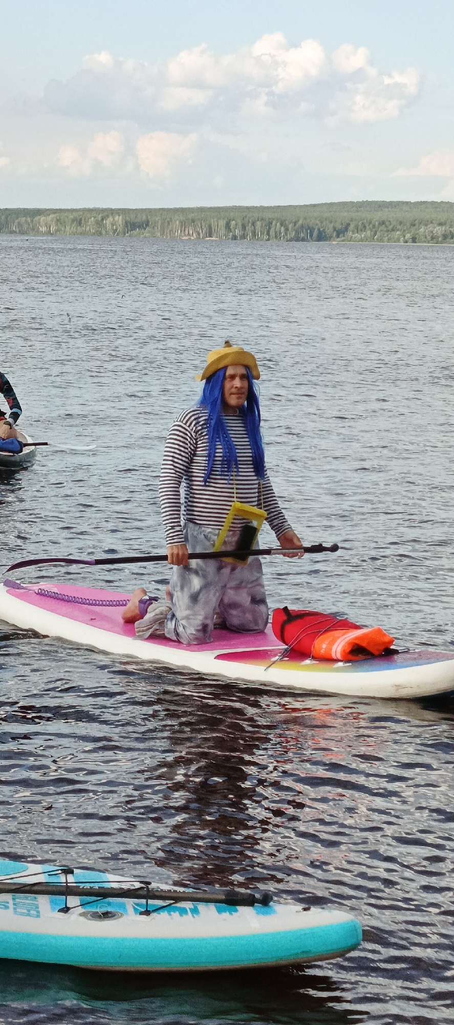 A man is kneeling on a pink and white paddleboard in a body of water, possibly a lake or river, with a forested shoreline in the background. He is wearing a yellow hat, a striped long-sleeved shirt, and patterned pants. A bright blue wig is visible under his hat, and a yellow bag containing a phone is hanging around his neck. He is holding a paddle and is positioned towards the front of the paddleboard, which also has a red life vest with orange straps strapped to it. Another paddleboard is partially visible in the foreground. The scene appears to be set during the day, with a cloudy sky. The overall impression is of a recreational activity, possibly part of a themed event or festival, given the man's costume. The location is identified as Chaikovsky, Russia.