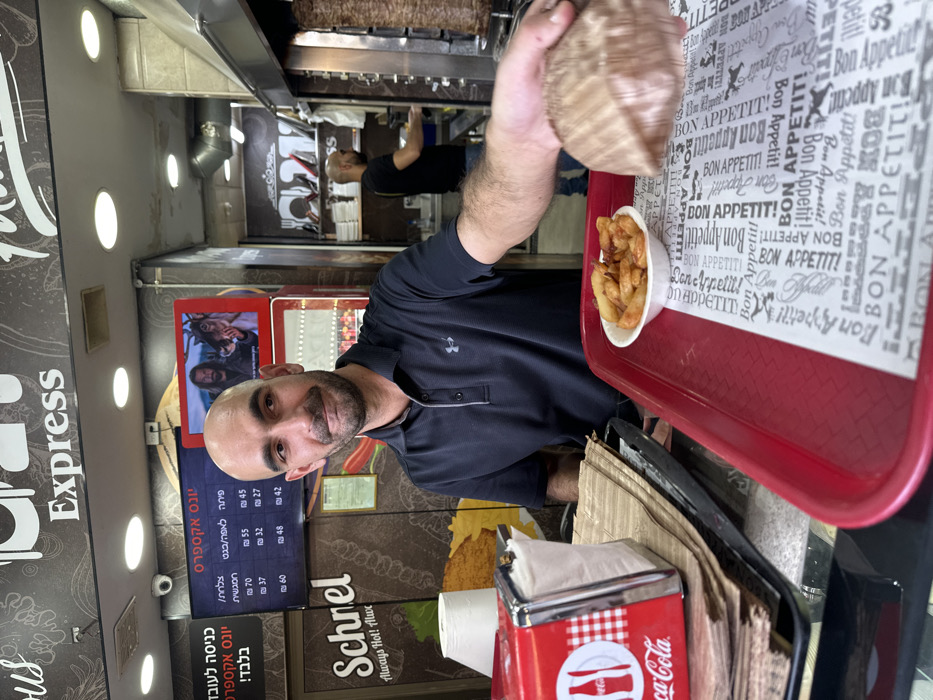 The image depicts the interior of a fast-food establishment named "Express" in Mishmar Hasharon, Israel. A bald, light-skinned adult male with a dark beard, wearing a dark polo shirt with an Under Armour logo, occupies the central foreground, smiling faintly and extending his right arm forward. His hand holds a wrapped food item, likely a sandwich or wrap, over a red plastic serving tray. The tray also holds a small white bowl filled with golden-brown french fries, and is lined with white paper displaying repeated "Bon Appetit!" text. To the left, a red and white checkered Coca-Cola napkin dispenser rests on a stack of brown paper bags/wrappers. In the background, behind the primary individual, a large digital menu board displays Hebrew text and prices (e.g., 70, 55, 45 ILS), alongside a smaller red-framed screen showing a video of a man with long hair and a deer. Further to the right in the background, another bald adult male, wearing a dark t-shirt, is partially visible operating a vertical rotisserie (shawarma spit) with meat. The ceiling is white with recessed lighting, and a small black security camera is mounted near the ceiling. Wall graphics include "Schnel Always Hot! Always Fresh!" and additional Hebrew text.