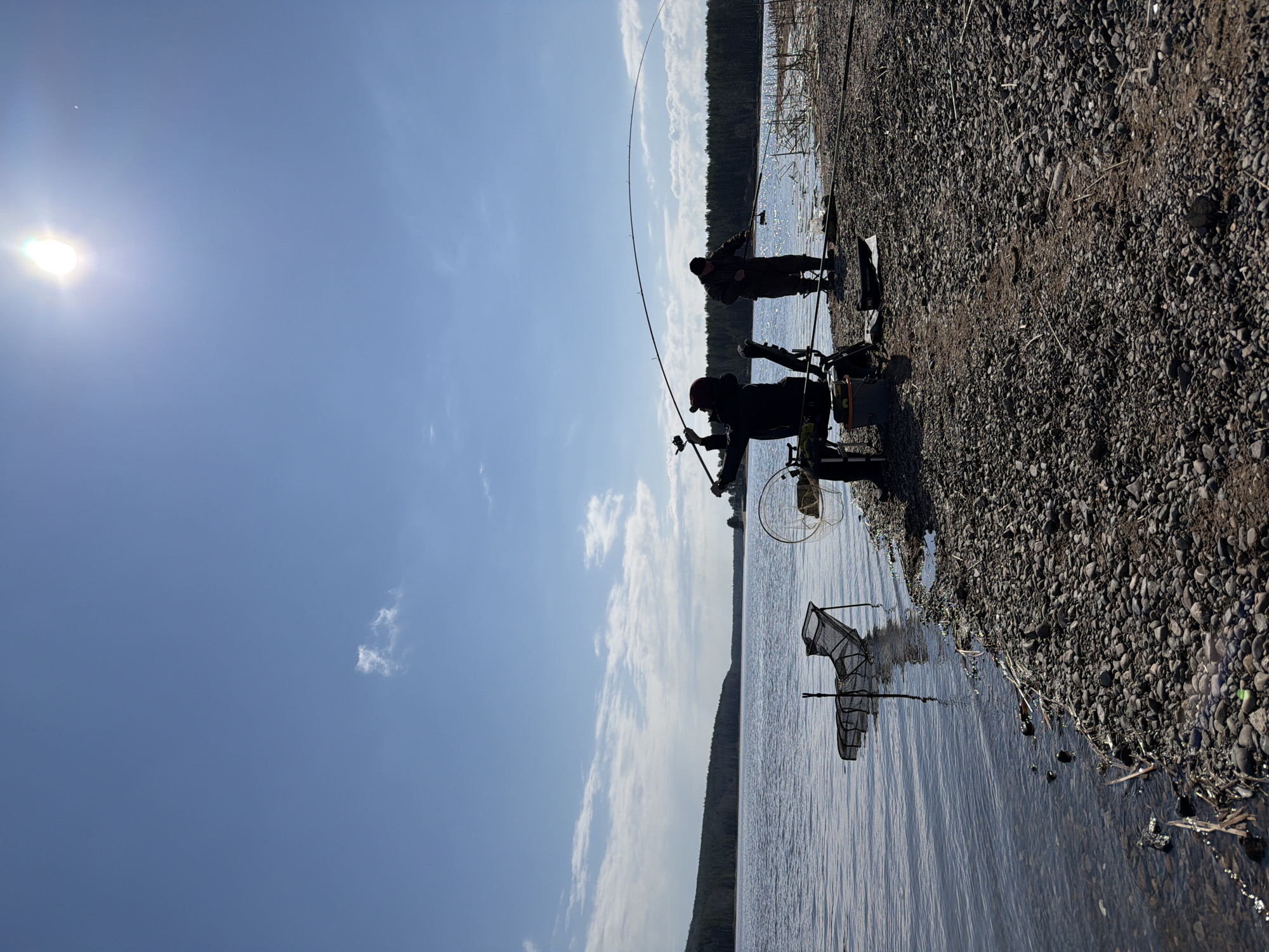 The image depicts two individuals fishing on a rocky shoreline of a body of water, possibly a lake or river. The scene is silhouetted against a bright, blue sky with scattered clouds and a visible sun. The shoreline consists of small pebbles and rocks. Fishing equipment, including fishing rods, nets, and a possible chair or fishing station, is visible along the shore. One individual is actively holding a fishing rod. The image orientation is landscape.
Specific location within Chaikovsky, Russia cannot be determined from the image.