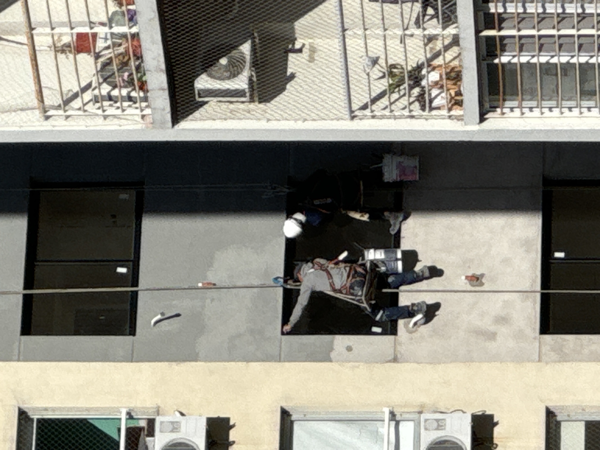 Aerial view of a building in Buenos Aires, Argentina. A construction worker in safety gear is rappelling down the building facade for repair work. The building is multiple stories, and some windows have bars. There is an air conditioning unit on the roof. Other appliances are also on the lower level.