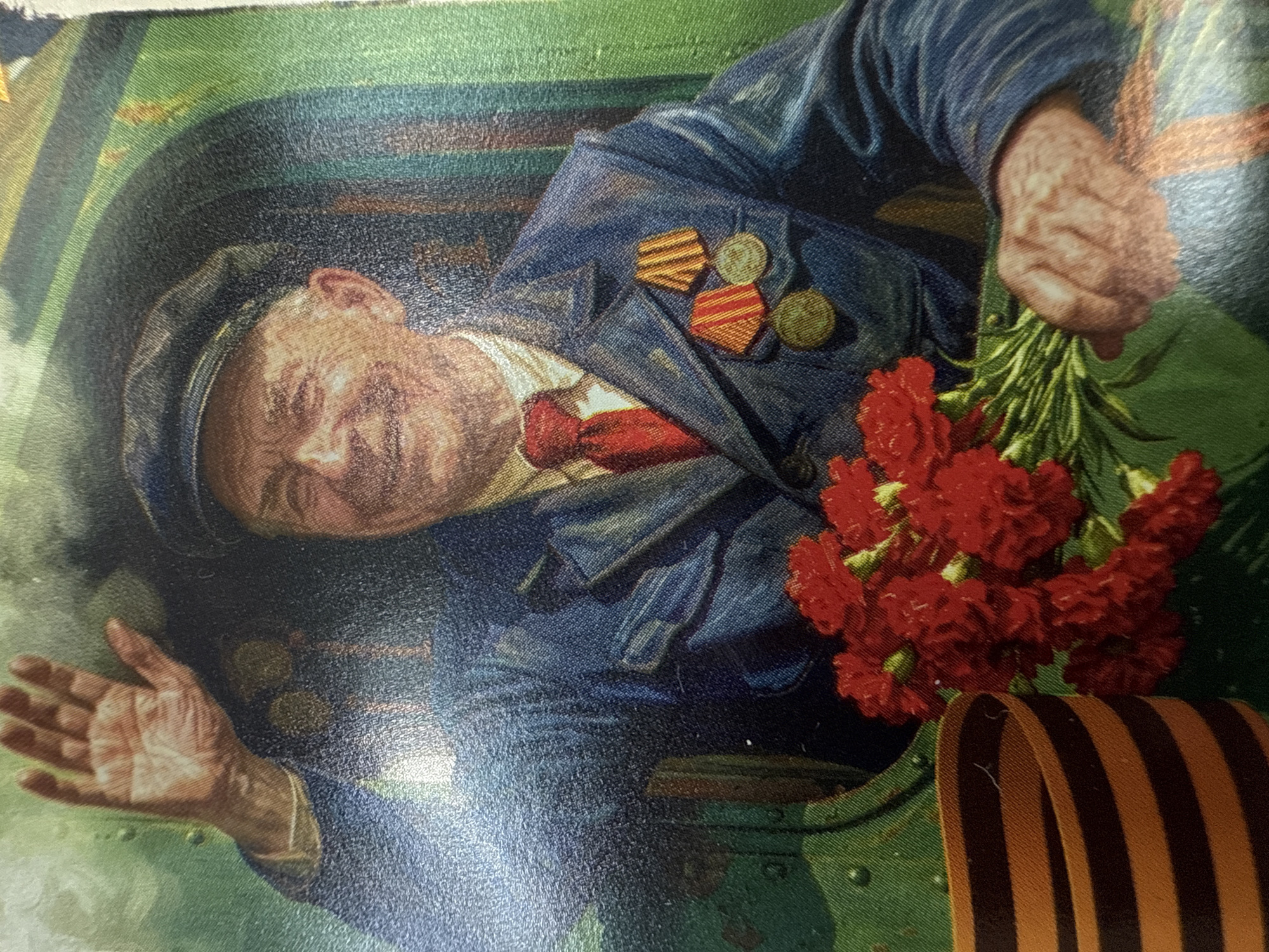 The image is a painting or print, presented here rotated roughly 90 degrees counter-clockwise. It depicts an older man, likely a veteran, from the chest up, against a dark, industrial-looking background.
The man has a fair complexion, short, light-colored hair, and a cap. His face shows signs of age, with a gentle, possibly reflective or proud expression. He wears a dark blue jacket with light vertical stripes on the sleeves, a white collared shirt, and a red tie. On his left lapel, he prominently displays several military decorations: two rectangular ribbon bars (one with orange and black stripes, another with red and yellow stripes) and three round, metallic medals.
In his left hand, the man holds a vibrant bouquet of red carnations, traditionally associated with remembrance and celebration. His right hand is extended forward, palm open, in a gesture that could signify greeting, offering, or inviting.
The background is dark and appears to be an interior space, possibly a cabin or the interior of heavy machinery, rendered in shades of dark green and blue, with hints of brown and lighter green suggesting metallic surfaces or reflections. In the bottom right corner of the composition, a distinct black and orange striped St. George's Ribbon is visible, a symbol of Russian military valor and remembrance.
The artwork's style is realistic with visible texture, suggesting it's a printed reproduction on a textured surface. The combination of military honors, carnations, and the St. George's Ribbon strongly suggests the scene is a commemoration of a veteran, likely tied to Victory Day in Russia, with the provided context placing this within Votkinsk, Russia. There is no visible text in the image.