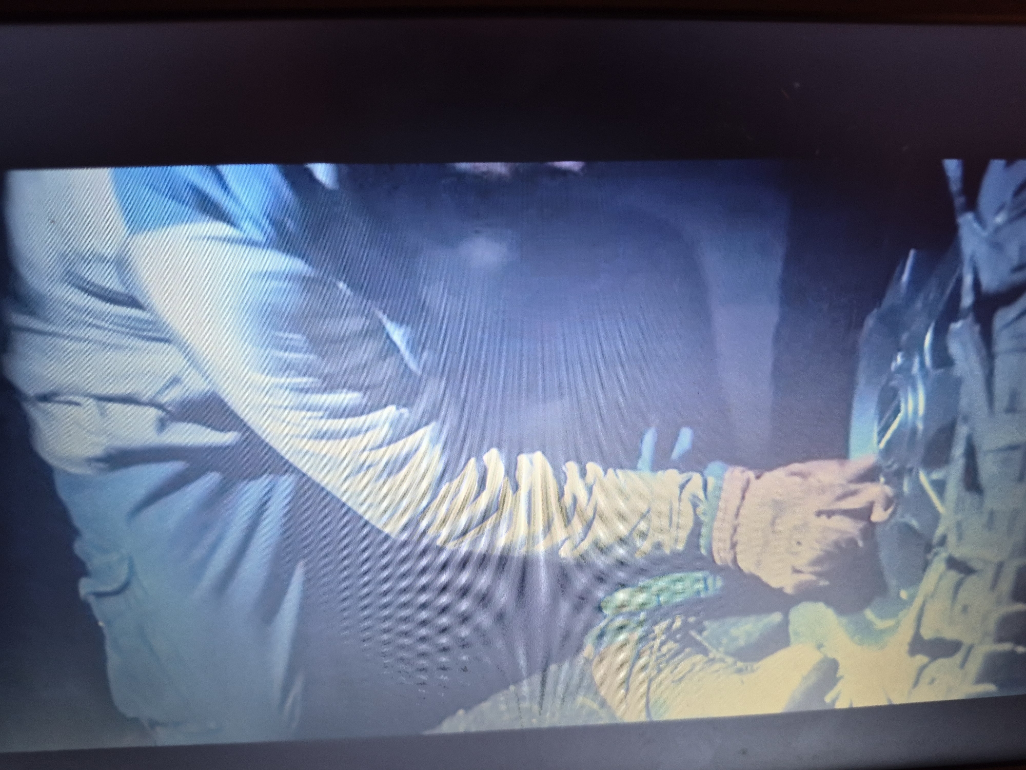 The image shows a close-up of two individuals' arms, appearing to be engaged in an activity in a low-light environment. The person on the left is wearing a light-colored long-sleeved shirt with blue accents on the shoulder and upper arm. Their right arm is bent, and the sleeve is ruched around the forearm. This individual is wearing a light-colored glove on their right hand, which is reaching towards the other person. The second person, on the right, is wearing a dark-colored garment, possibly a tactical vest or backpack, with straps and buckles visible. Their arm is also visible, and it appears they are holding or manipulating an object. The background is dark and indistinct, with some light reflecting off surfaces, suggesting an indoor or nighttime outdoor setting. There is no discernible information in the image to indicate a specific location within the city of Sandwell, United Kingdom.