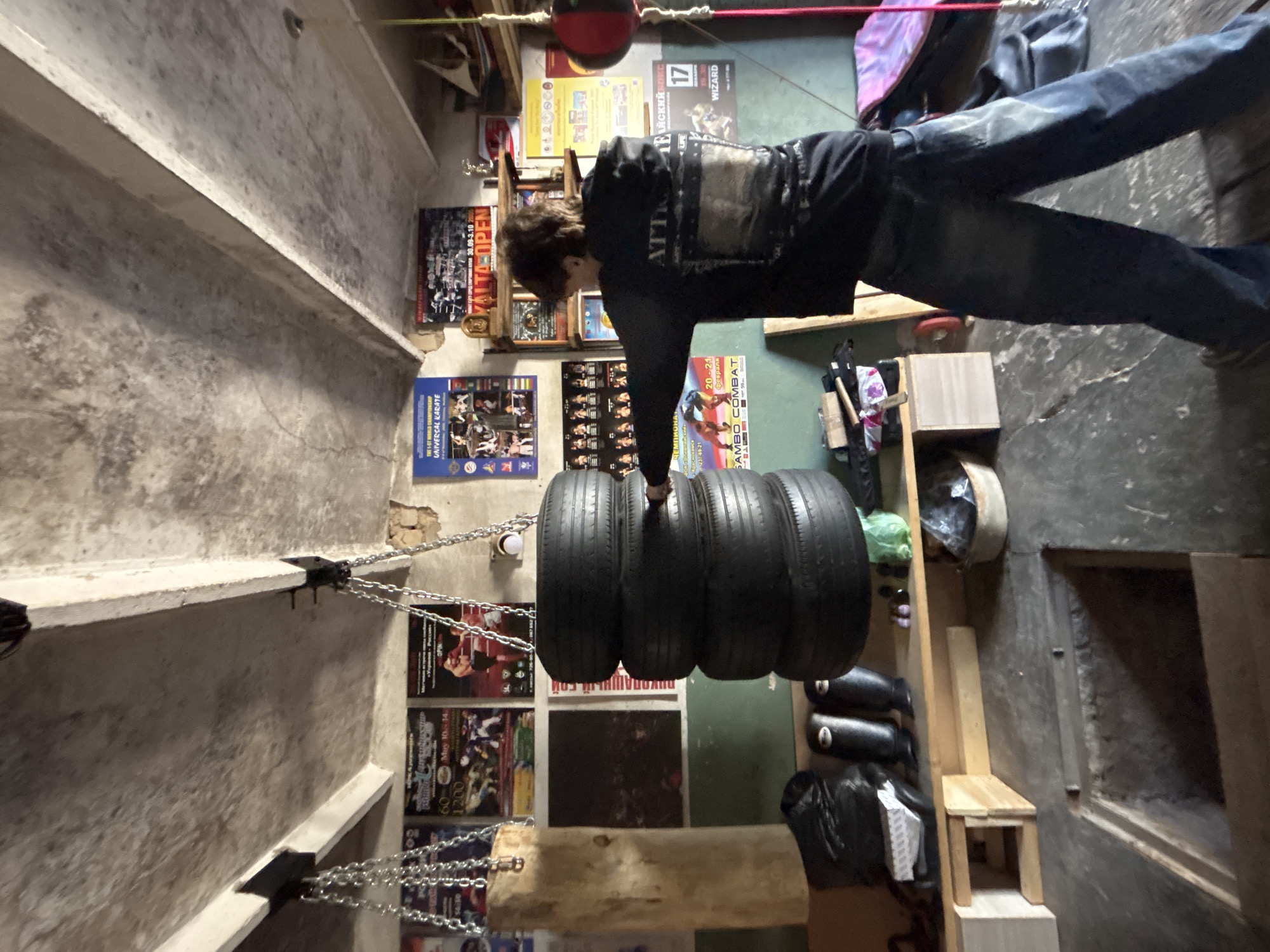 The image captures a scene inside a boxing or martial arts training area. A person is seen standing and reaching towards a hanging punching bag constructed from tires. The walls are decorated with various posters promoting combat sports events, some written in Russian. Text visible in the images indicates a location in Krasnokamsk, Russia, and mentions the city of Chaikovsky. The training area appears to be located in an indoor concrete building. There are additional items such as boots, equipment bags, wooden planks, and shelves stored nearby.